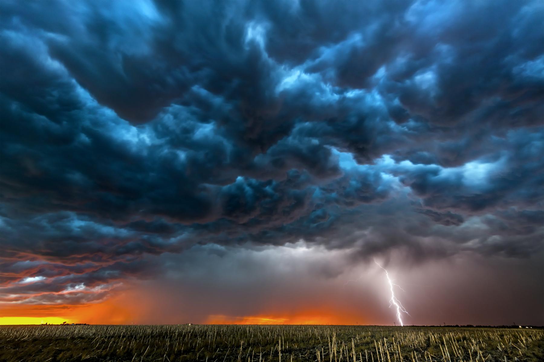 Gewitterwolken über einem Feld bei Sonnenuntergang mit einem Blitzschlag am Horizont