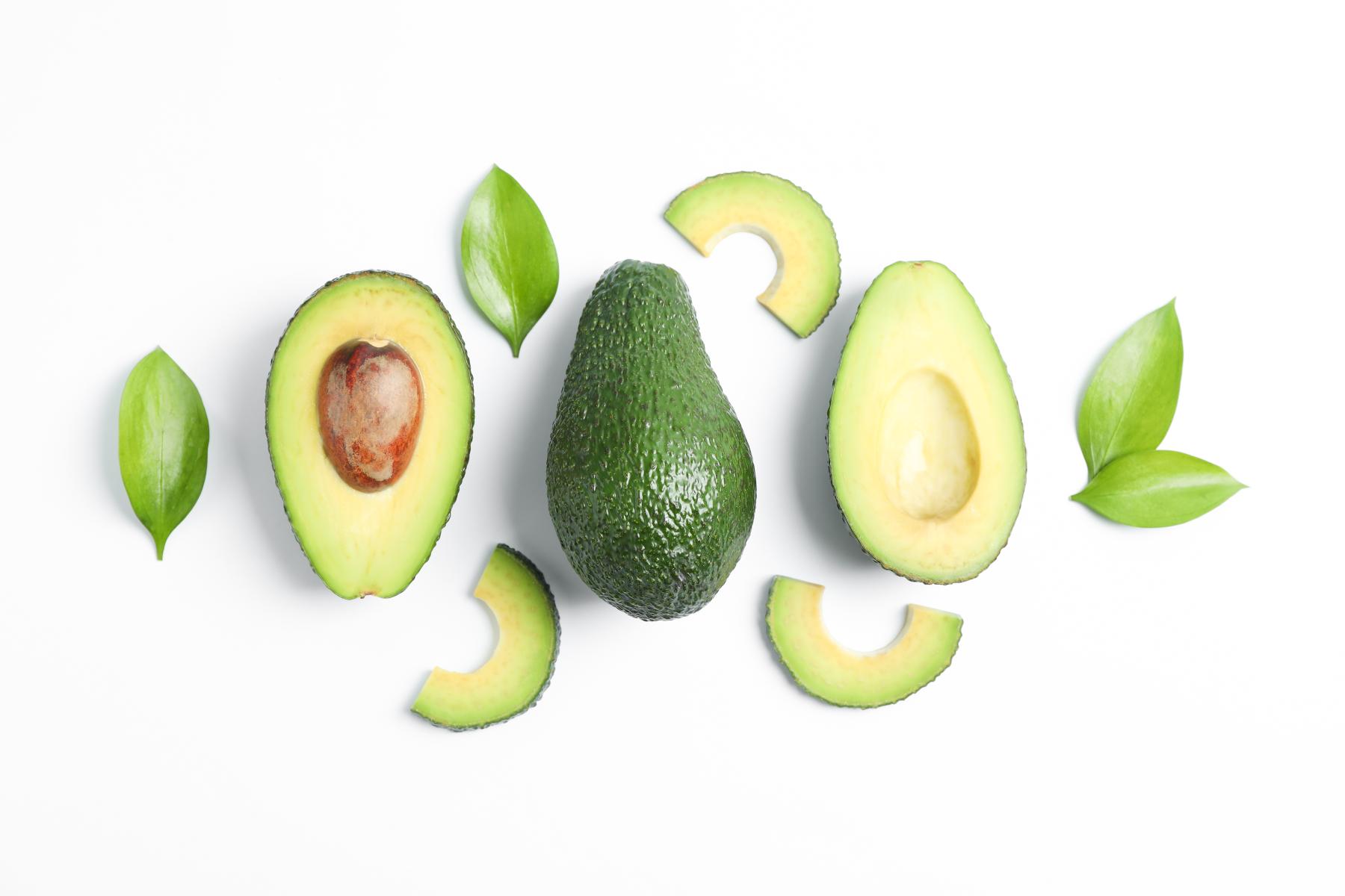 Ganze und halbierte Avocado mit Kern und Scheiben, umgeben von grünen Blättern auf weißem Hintergrund