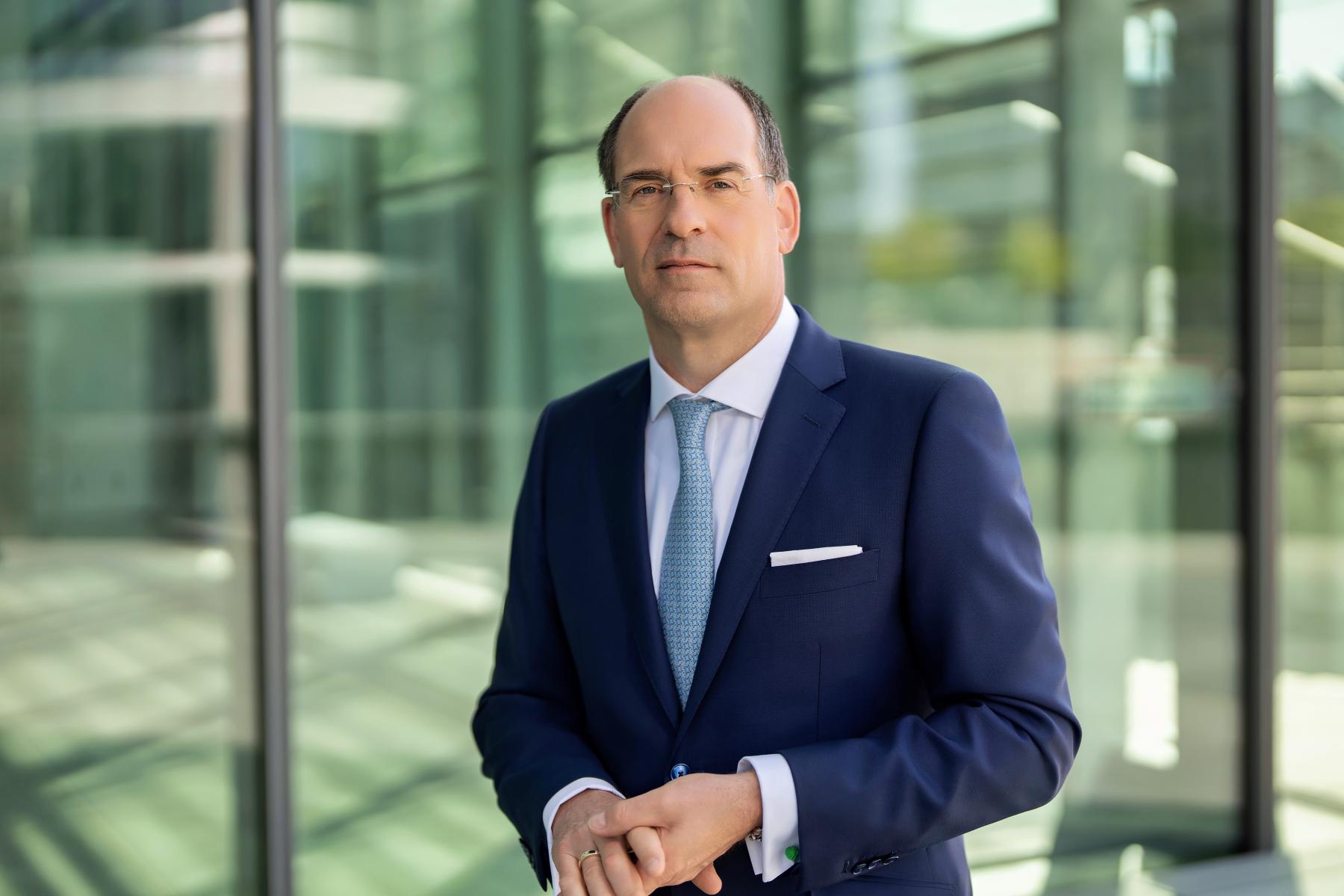 BGA-Präsident Dr. Dirk Jandura steht in einem modernen Glasgebäude, trägt einen dunkelblauen Anzug mit weißem Hemd und blauer Krawatte