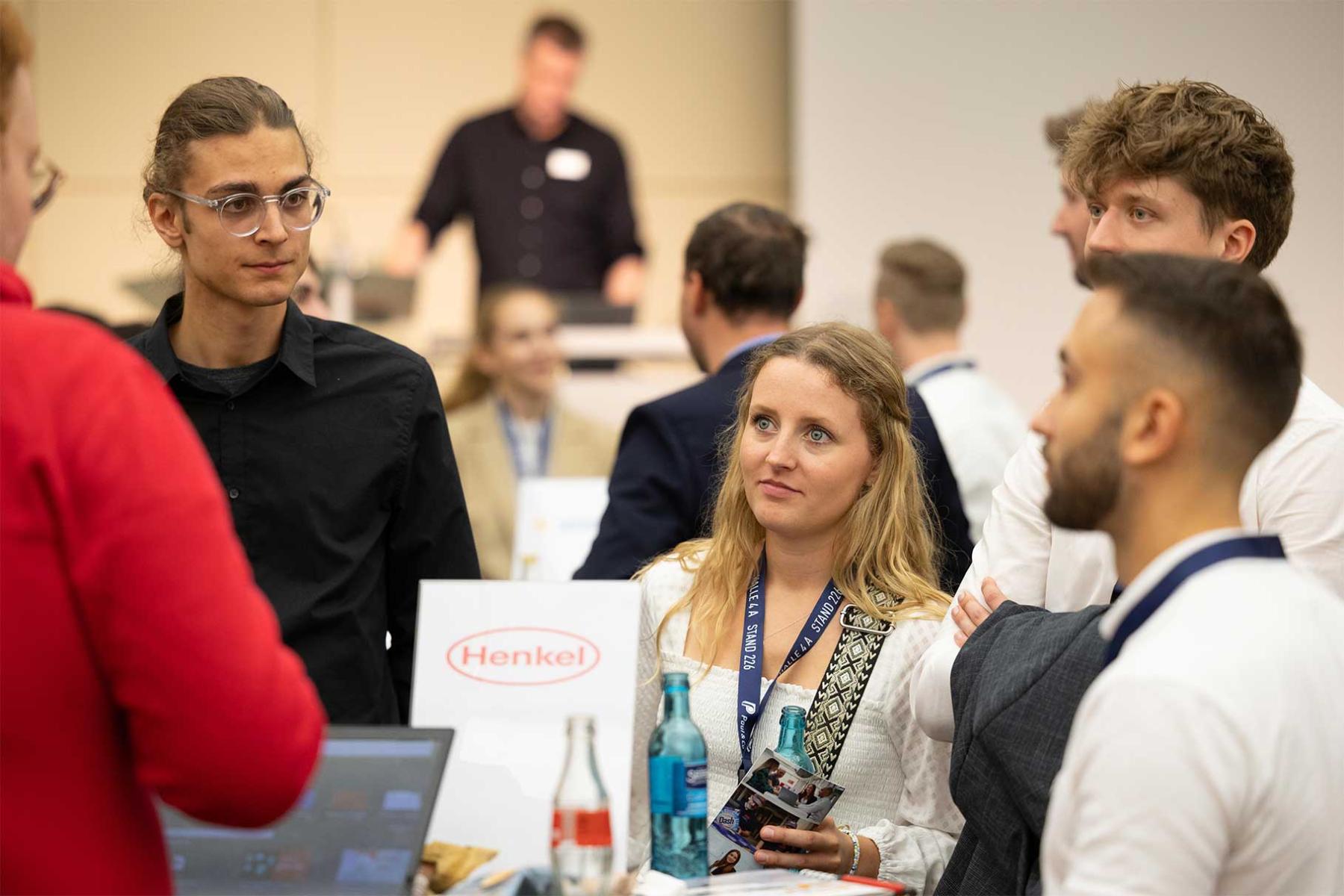 Mehrere Personen stehen und unterhalten sich auf einer Messe, im Vordergrund ein Tisch mit Henkel-Logo und Getränken