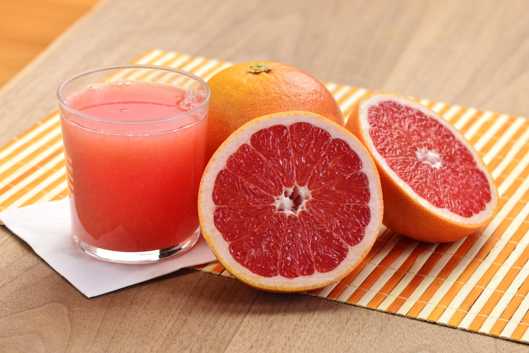 Frisch gepresster Grapefruitsaft im Glas neben einer ganzen und zwei halbierten Grapefruits auf einem gestreiften Untersetzer