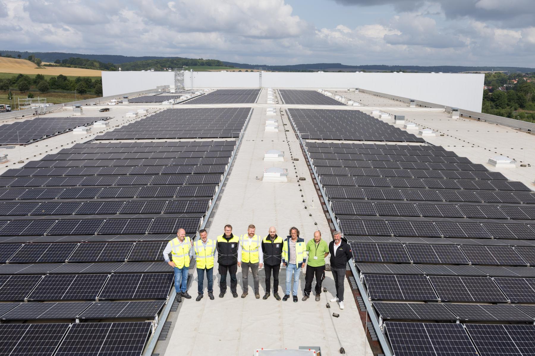 Acht Personen in Sicherheitswesten stehen auf einem Flachdach zwischen großen Solarpanelreihen unter bewölktem Himmel
