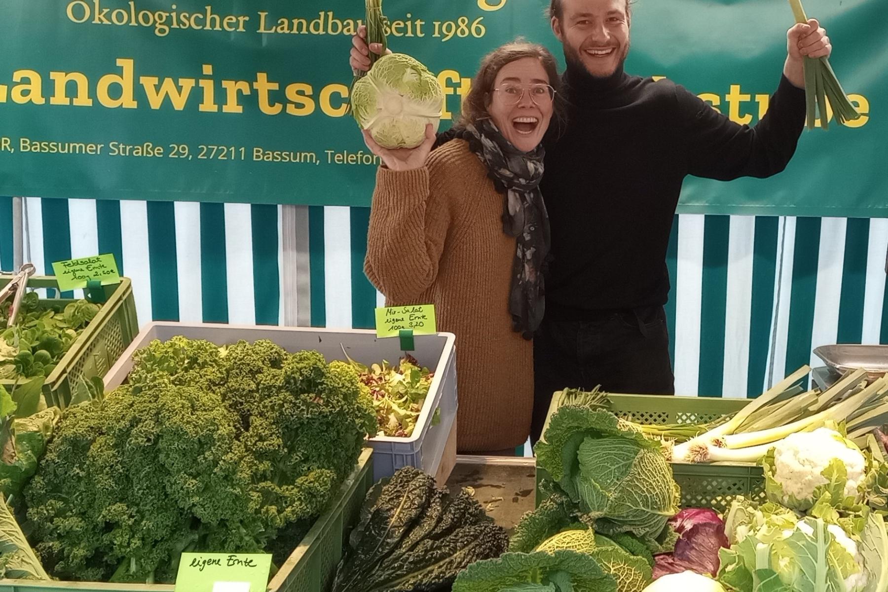 Julia Albrecht und Mitarbeiter Nicolas Hasselbeck stehen hinter einem Marktstand mit frischem Bio-Gemüse wie Grünkohl, Kohlrabi und Lauch, Banner 'Biolandhof Steding Ökologischer Landbau seit 1986' im Hintergrund