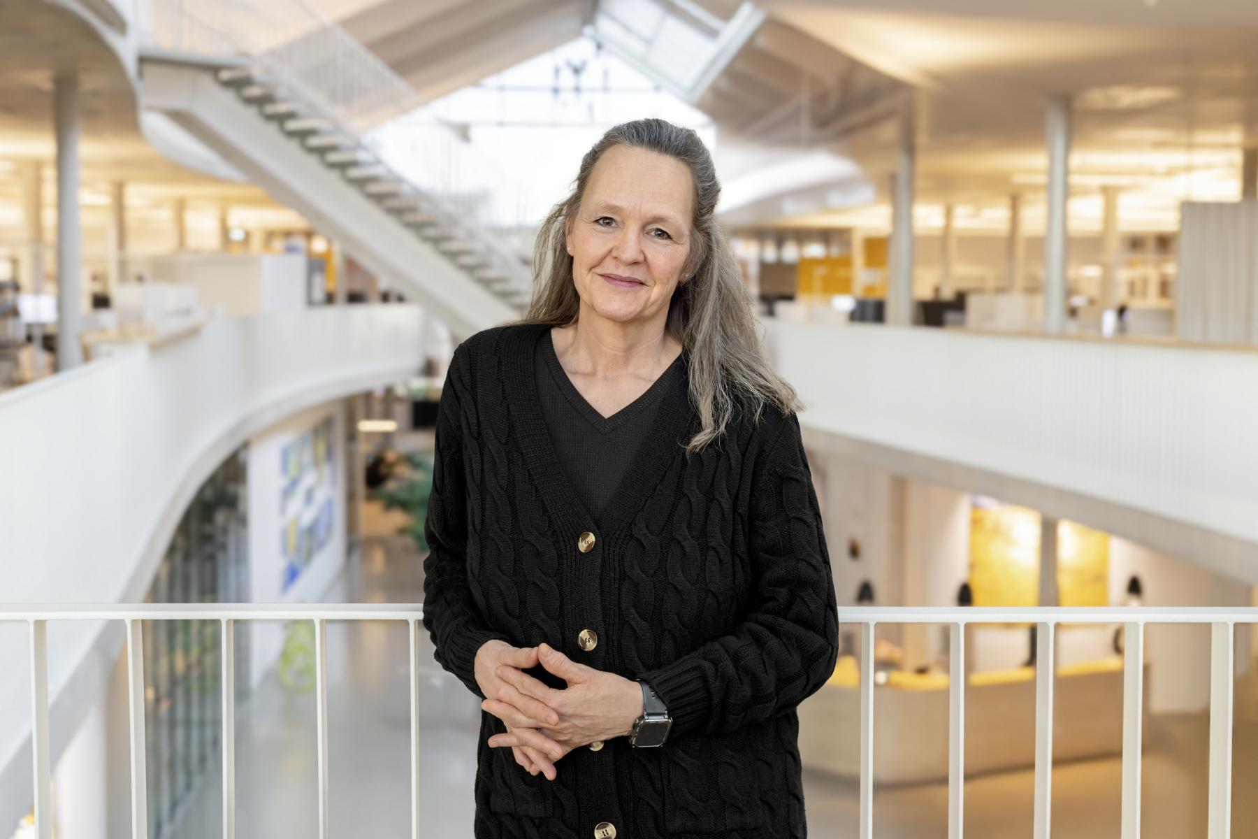 Kirsten Keller steht in einem modernen Bürogebäude mit hellem Innenraum, trägt einen schwarzen Strickcardigan und eine Smartwatch