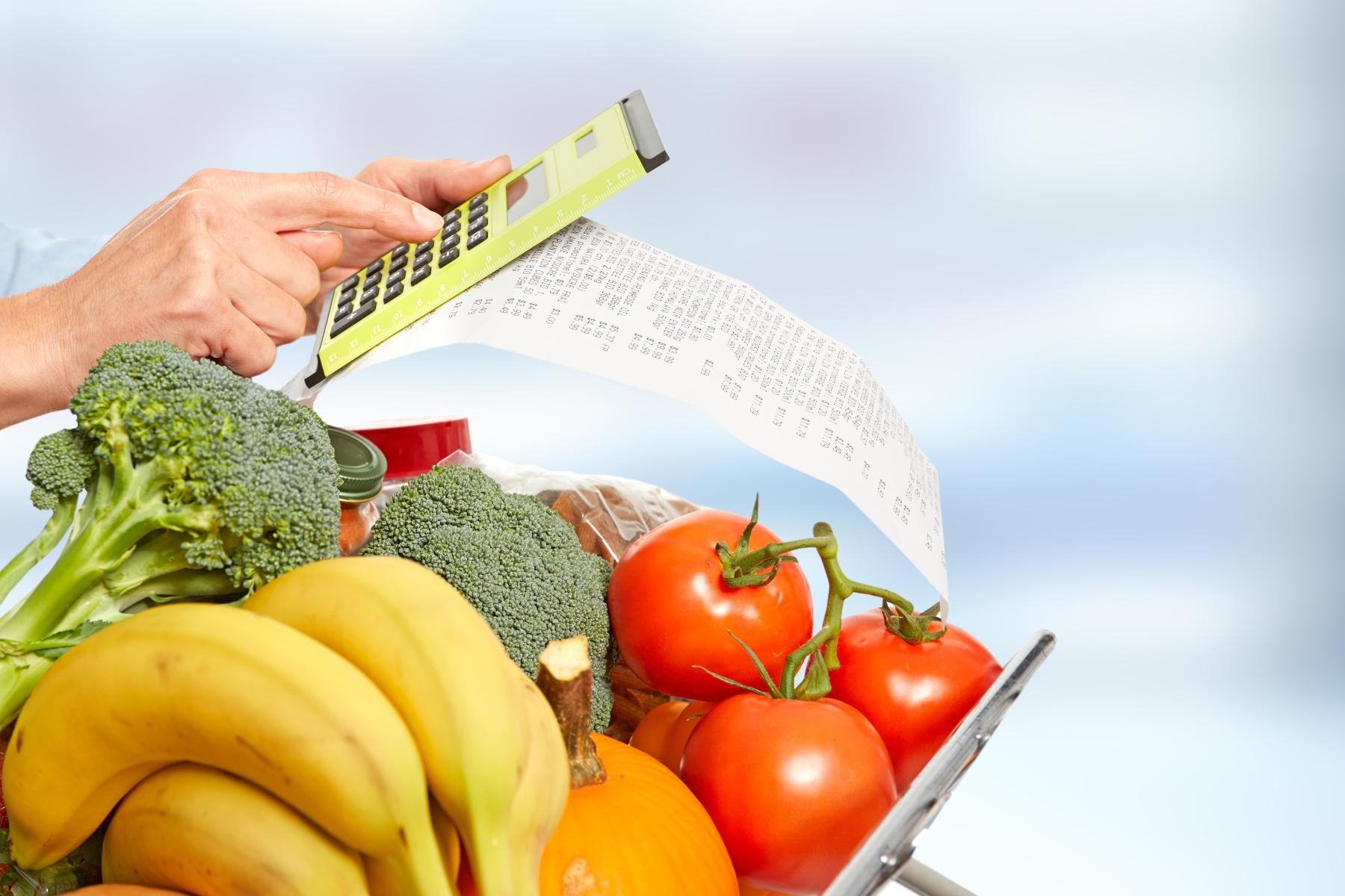 Person berechnet mit Taschenrechner die Kosten eines Einkaufs mit frischem Gemüse und Obst wie Brokkoli, Bananen und Tomaten