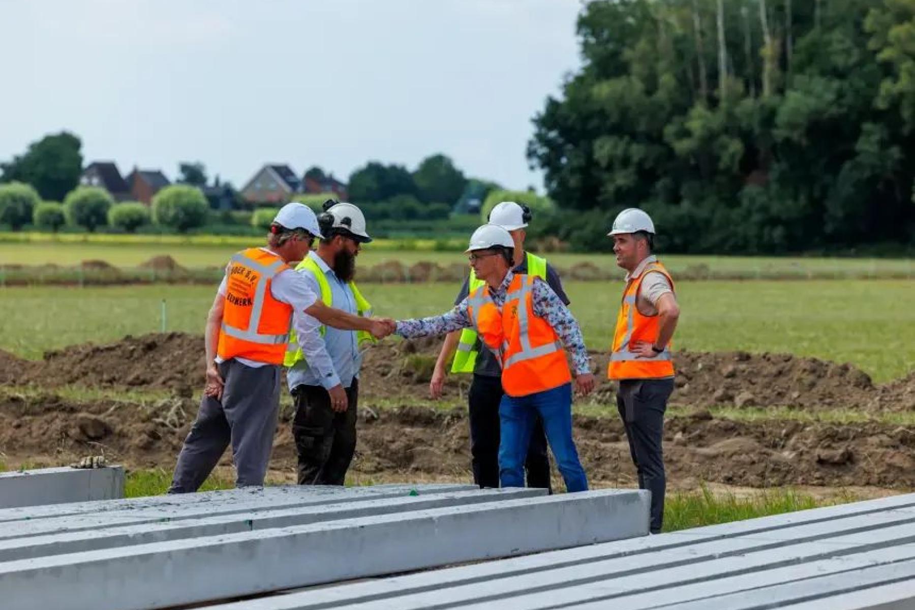 Fünf Personen auf der Baustelle. Sie tragen Schutzhelme und orange Warnwesten, zwei schütteln sich die Hand