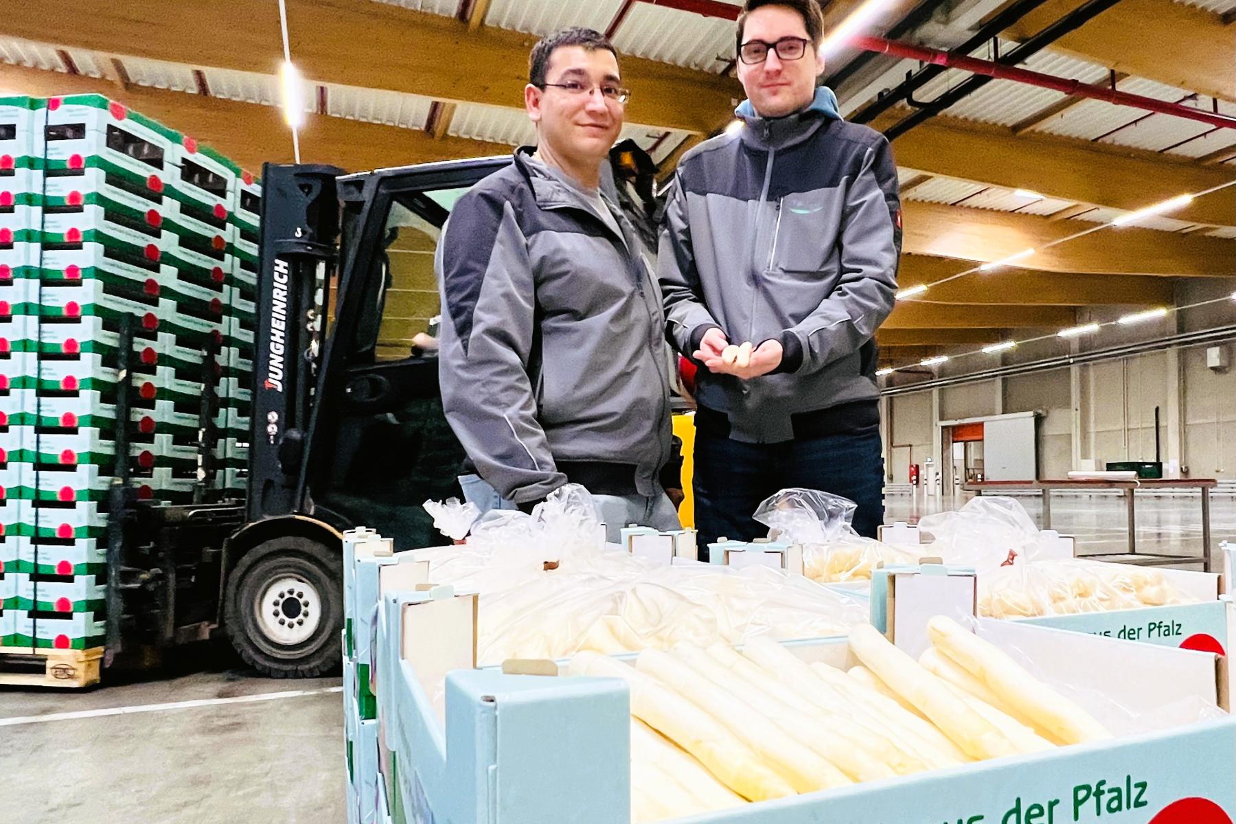 Hugo da Motto und Leo Zastrow vom Spargelbüro der Pfalzmarkt eG stehen in einer Lagerhalle neben Kisten mit frischem Spargel aus der Pfalz