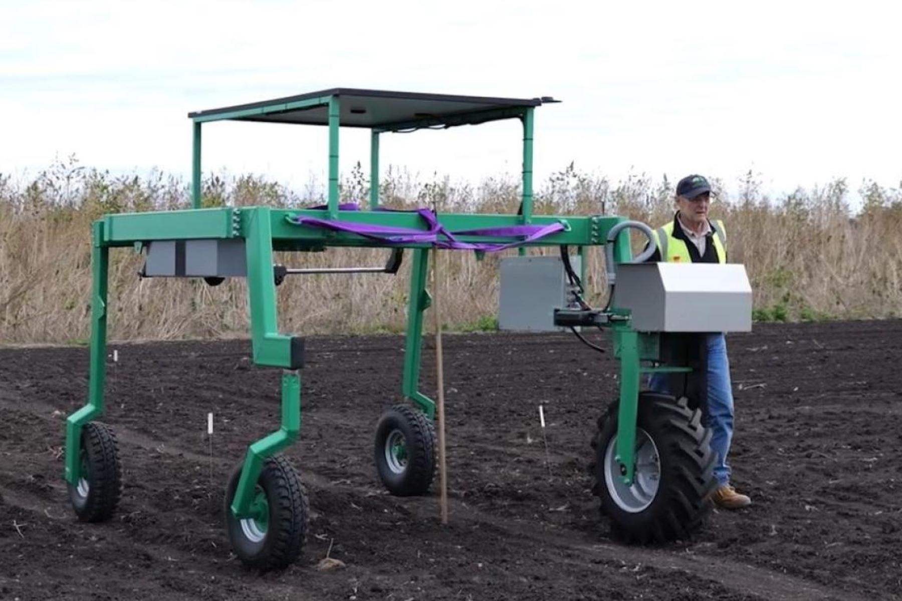Prof. Daniel Rodriguez steht neben dem RootBot, einem großen grünen landwirtschaftlichen Roboter mit vier Rädern auf einem Acker