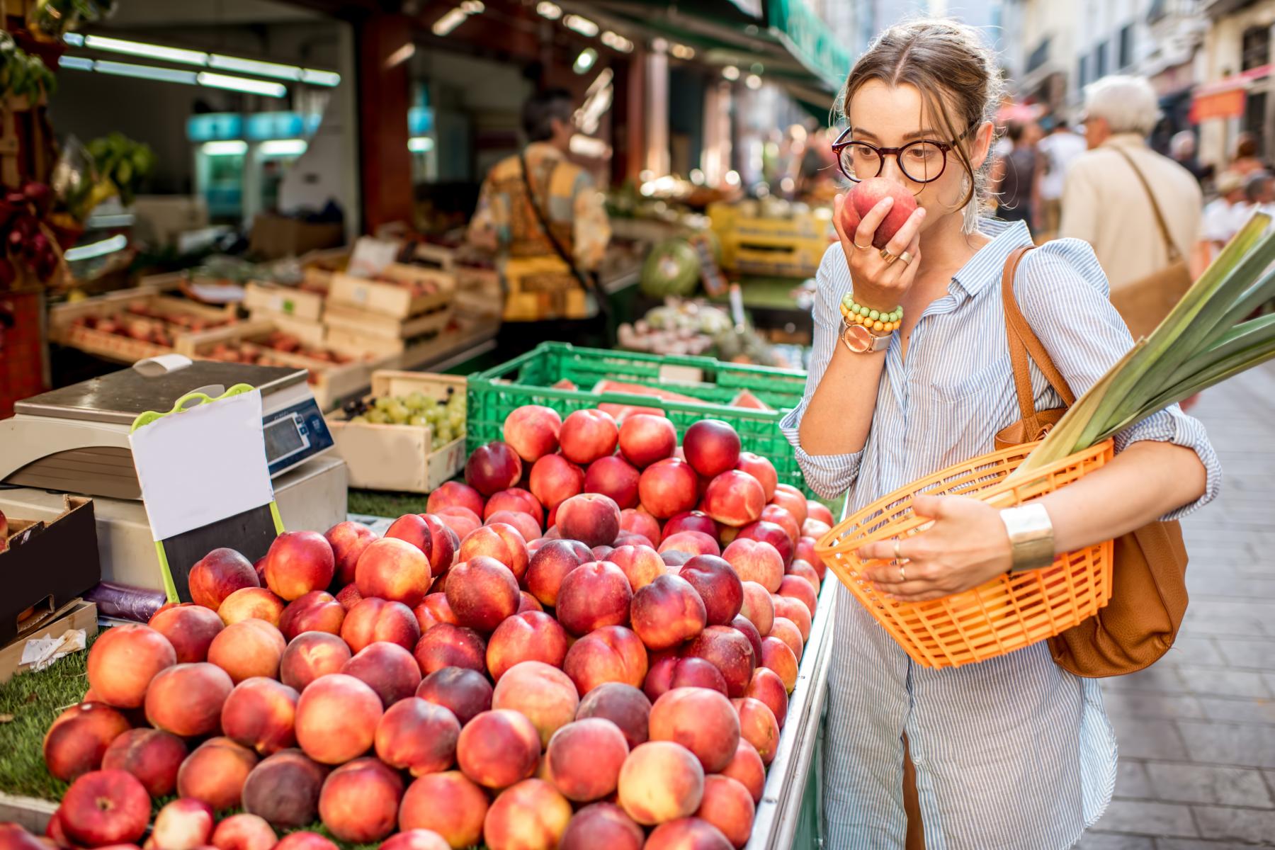 Frau hält Einkaufskorb mit Lauch in der einen und einen Pfirsich in der anderen Hand. Sie steht an Marktstand mit Pfirsichen und anderem Obst