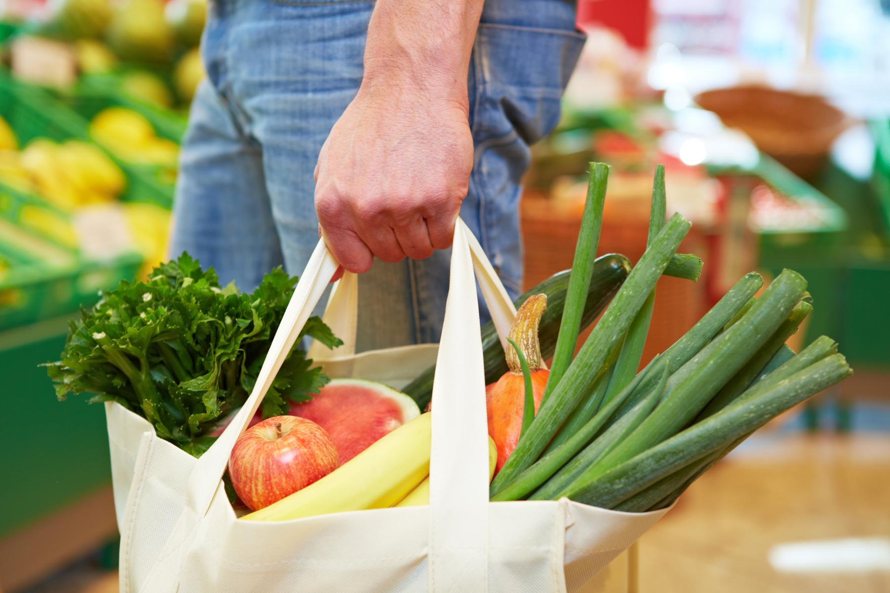 Hand hält eine Stofftasche mit frischem Gemüse und Obst wie Äpfeln, Bananen, Frühlingszwiebeln und Petersilie im Supermarkt