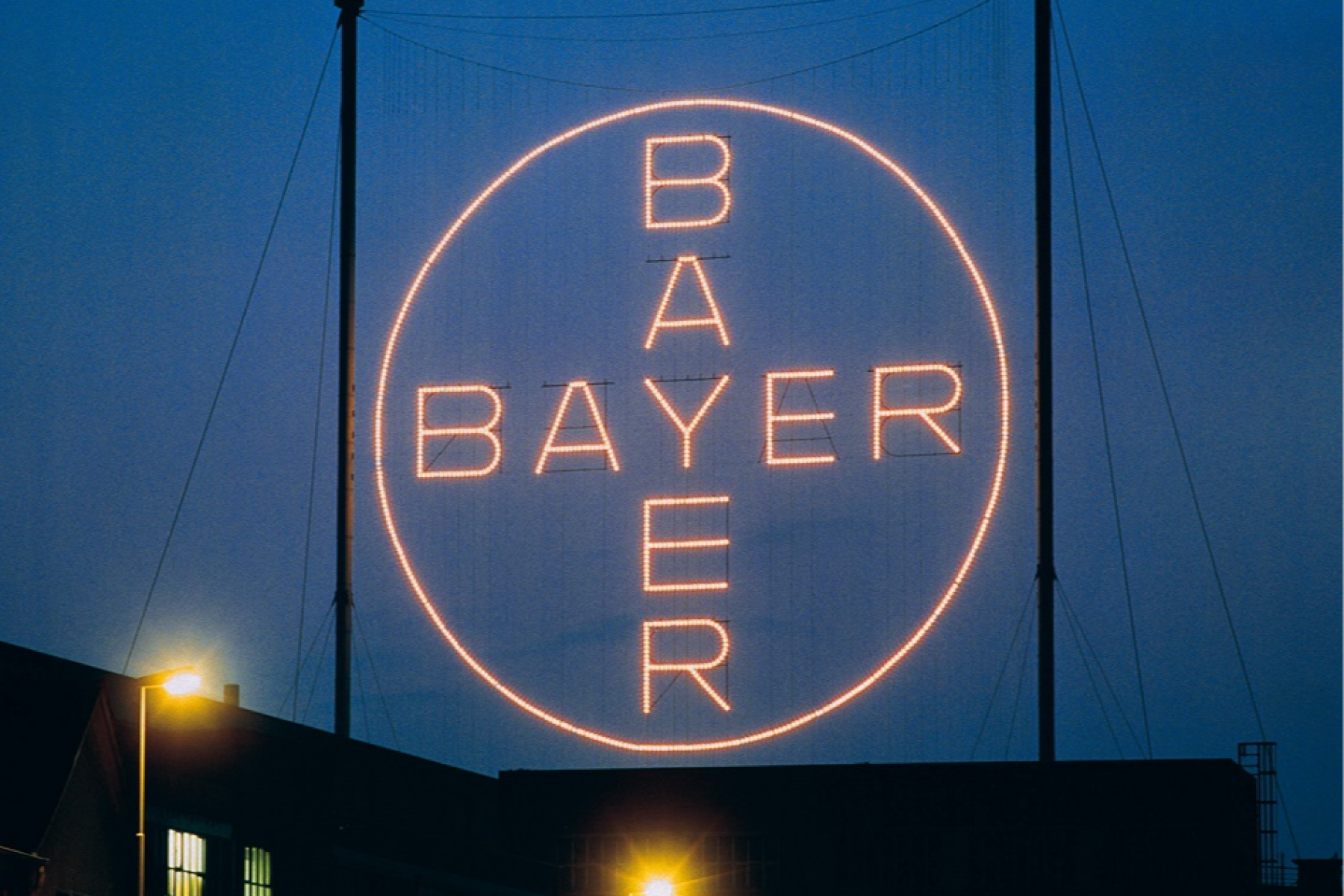Leuchtendes Bayer-Kreuz mit dem Schriftzug 'BAYER' in einem Kreis, nachts vor dunklem Himmel