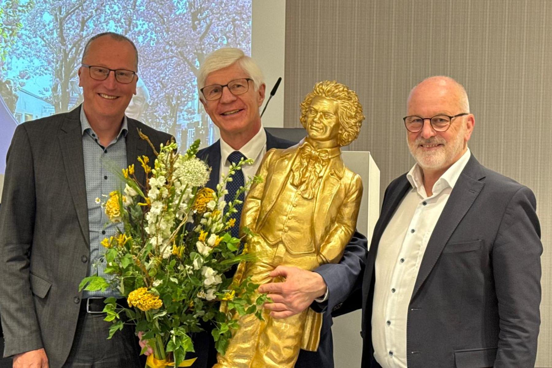 Jürgen Bruch, Dieter Krauß und Dr. Andreas Brügger stehen zusammen, halten eine goldene Statue und einen Blumenstrauß. Im Hintergrund ist eine Präsentation sichtbar.