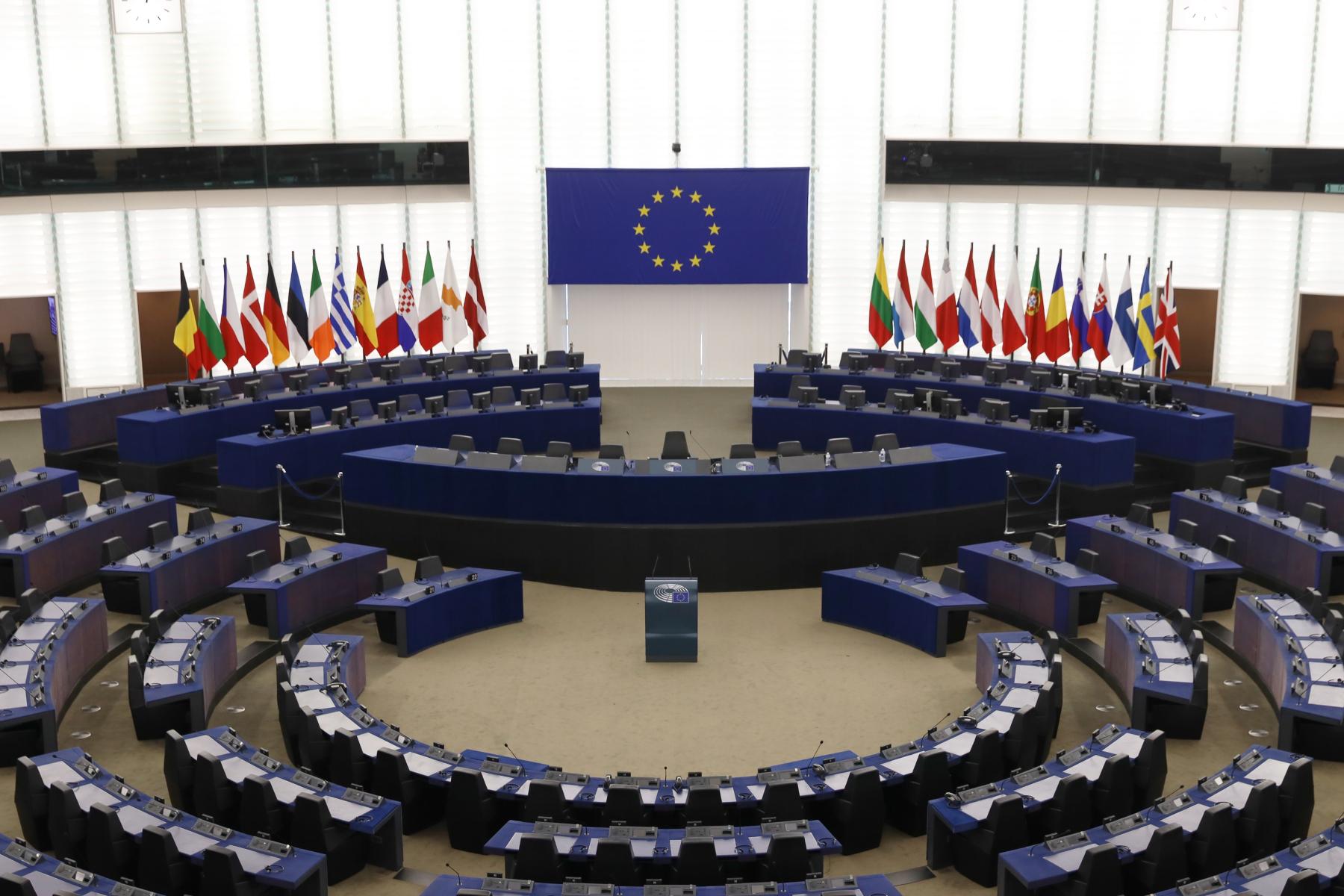 Leerer Plenarsaal des Europa Parlaments mit halbkreisförmigen Sitzreihen, Rednerpult und EU-Flagge an der Wand, flankiert von mehreren Nationalflaggen