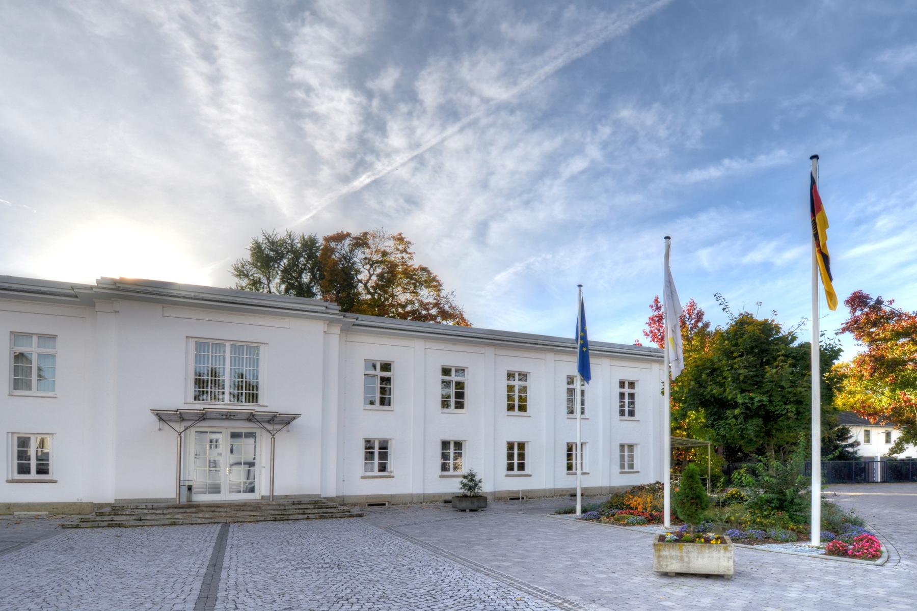 Gebäude des Bundeskartellamtes, weißes Verwaltungsgebäude mit mehreren Fenstern, drei Flaggenmasten mit EU-, Deutschland- und Bundeskartellamtsflagge vor dem Eingang