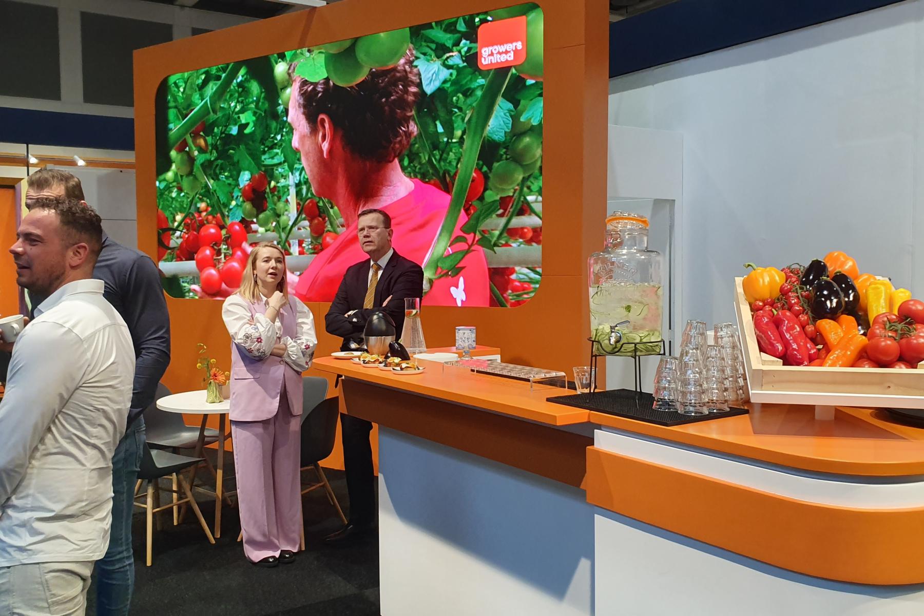 Messestand von Growers United mit großem Bildschirm, der eine Frau vor Tomaten zeigt, und einem Tresen mit frischem Gemüse und Getränkespender