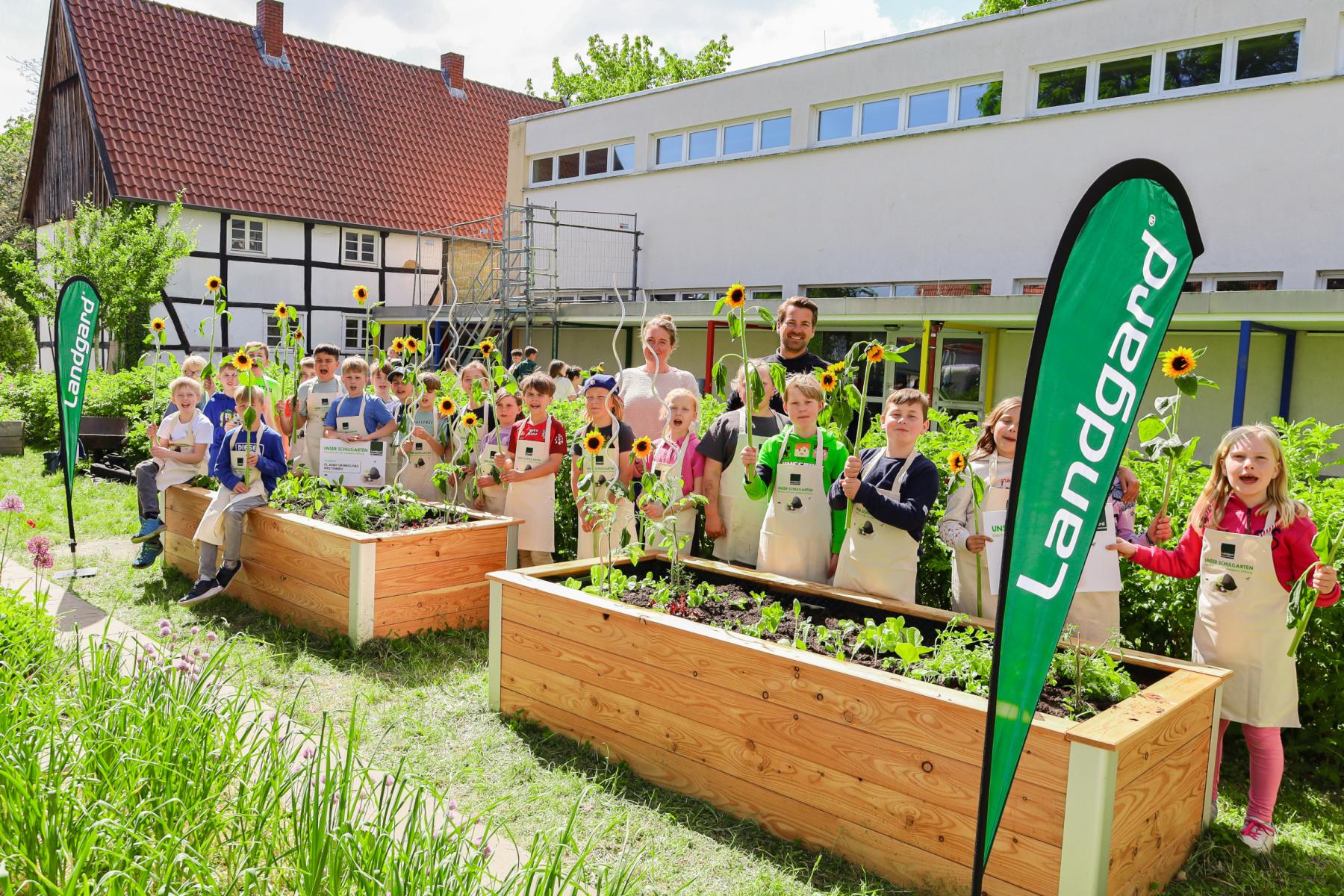 Gruppe von Kindern in Schürzen, die in einem Schulgarten stehen, umgeben von Hochbeeten mit Sonnenblumen und Gemüse. Zwei grüne Fahnen mit 'Landgard'
