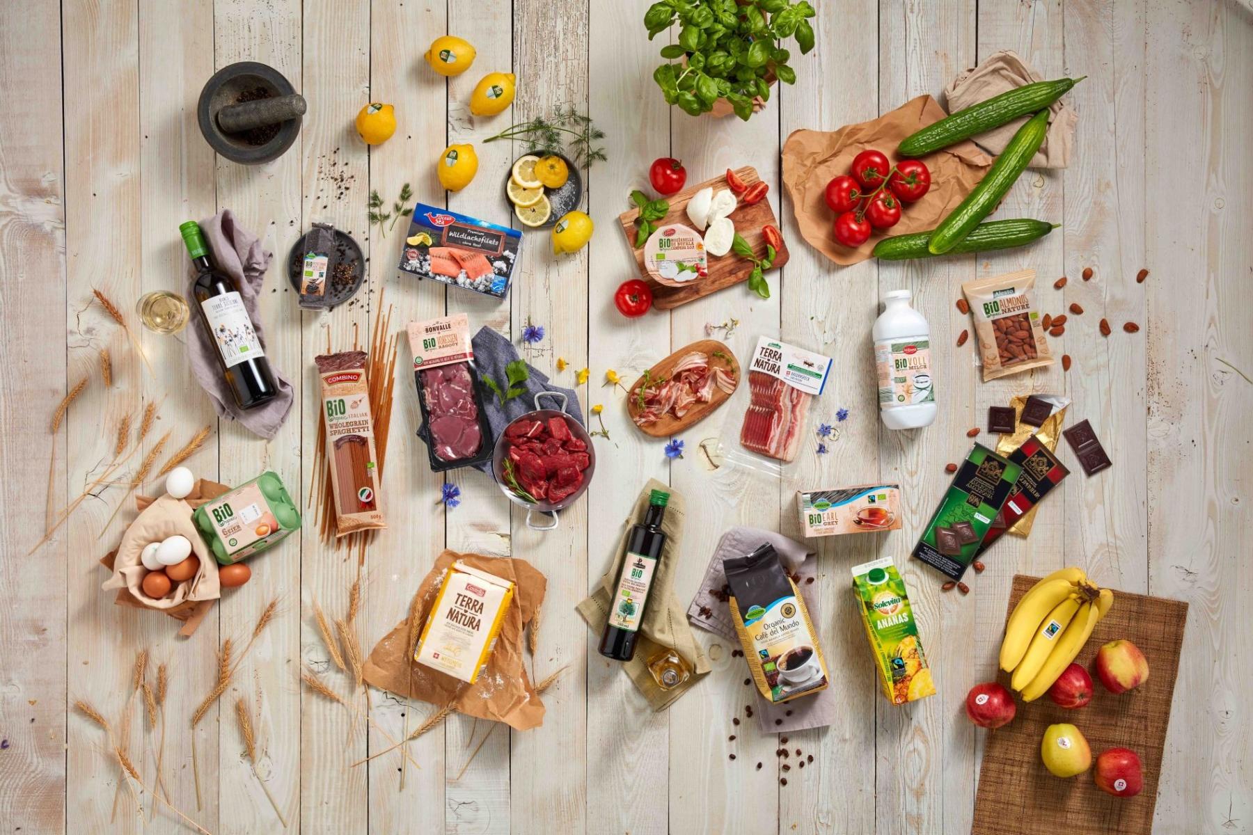 Verschiedene Lebensmittel auf hellem Holztisch, darunter Wein, Eier, Bio-Fleisch, Gemüse, Obst, Nudeln, Milchprodukte und Säfte, arrangiert in Gruppen.