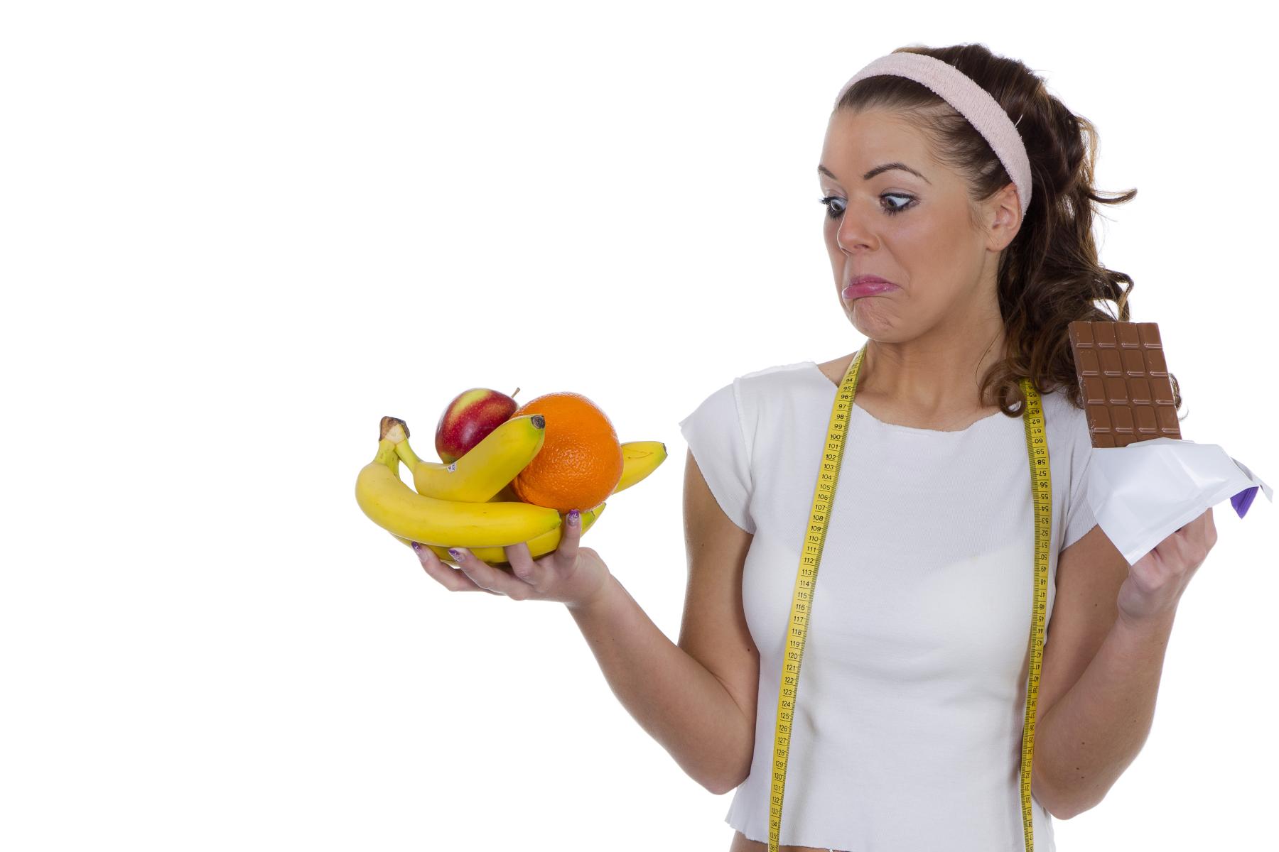 Frau mit Maßband um den Hals hält in einer Hand eine Schale mit Bananen, Apfel und Orange, in der anderen Hand eine Tafel Schokolade