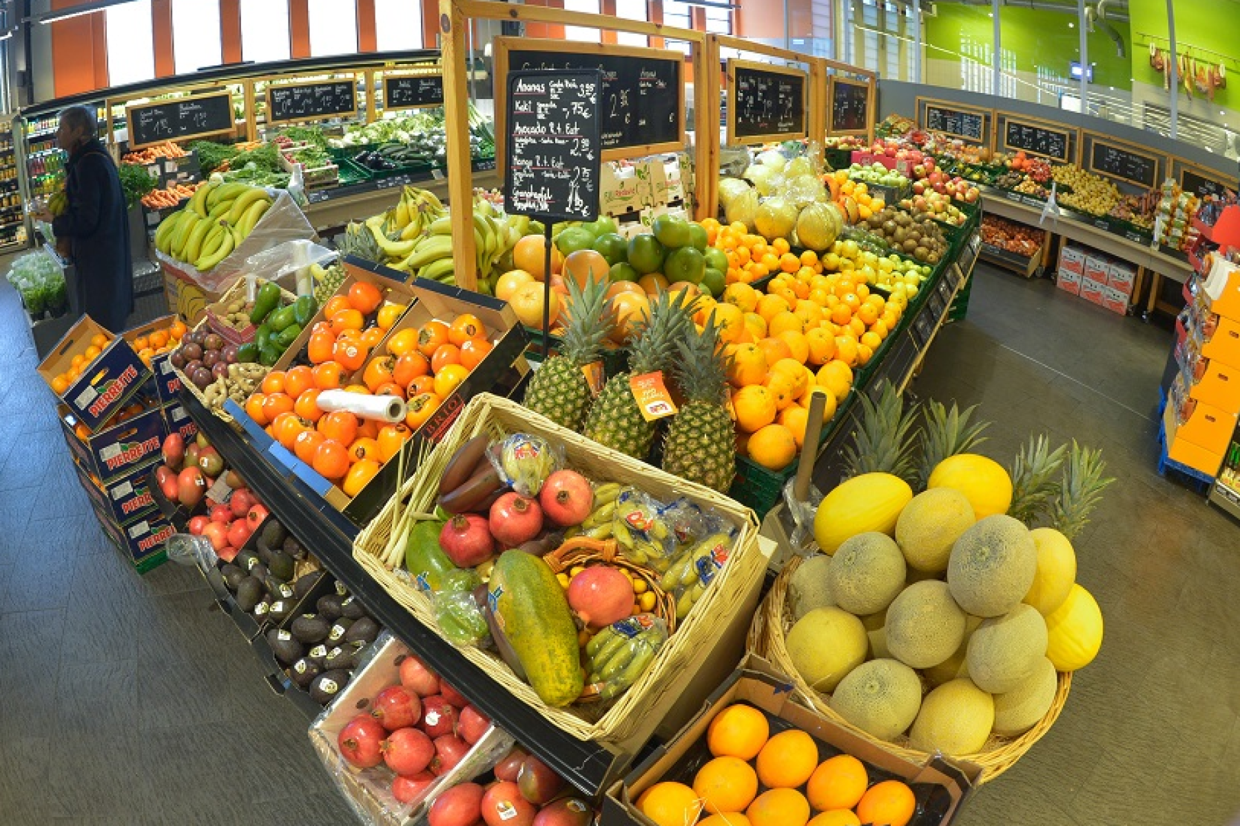 Obst- und Gemüseabteilung in einem Supermarkt mit verschiedenen Früchten wie Bananen, Ananas, Orangen, Melonen und Äpfeln auf Verkaufstischen