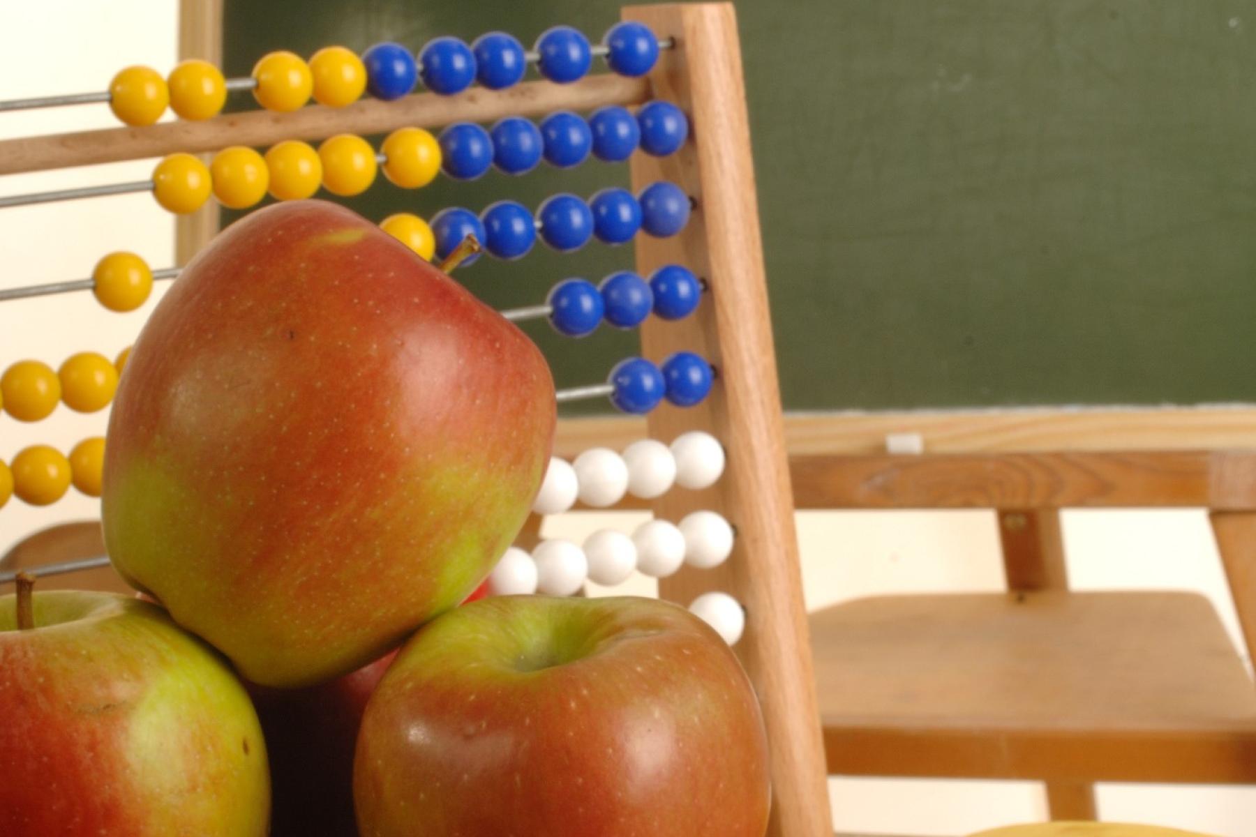 Drei rote Äpfel vor einem Abakus mit gelben, blauen und weißen Perlen in einem Klassenzimmer