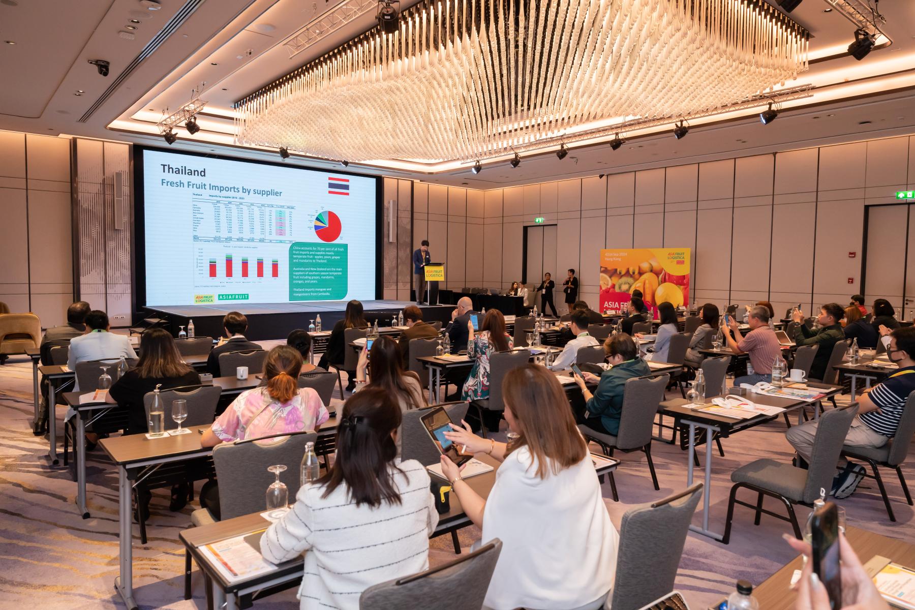 Konferenzraum mit Teilnehmern, die einer Präsentation über Frischobstimporte in Thailand folgen, Sprecher auf dem Podium vor großem Bildschirm