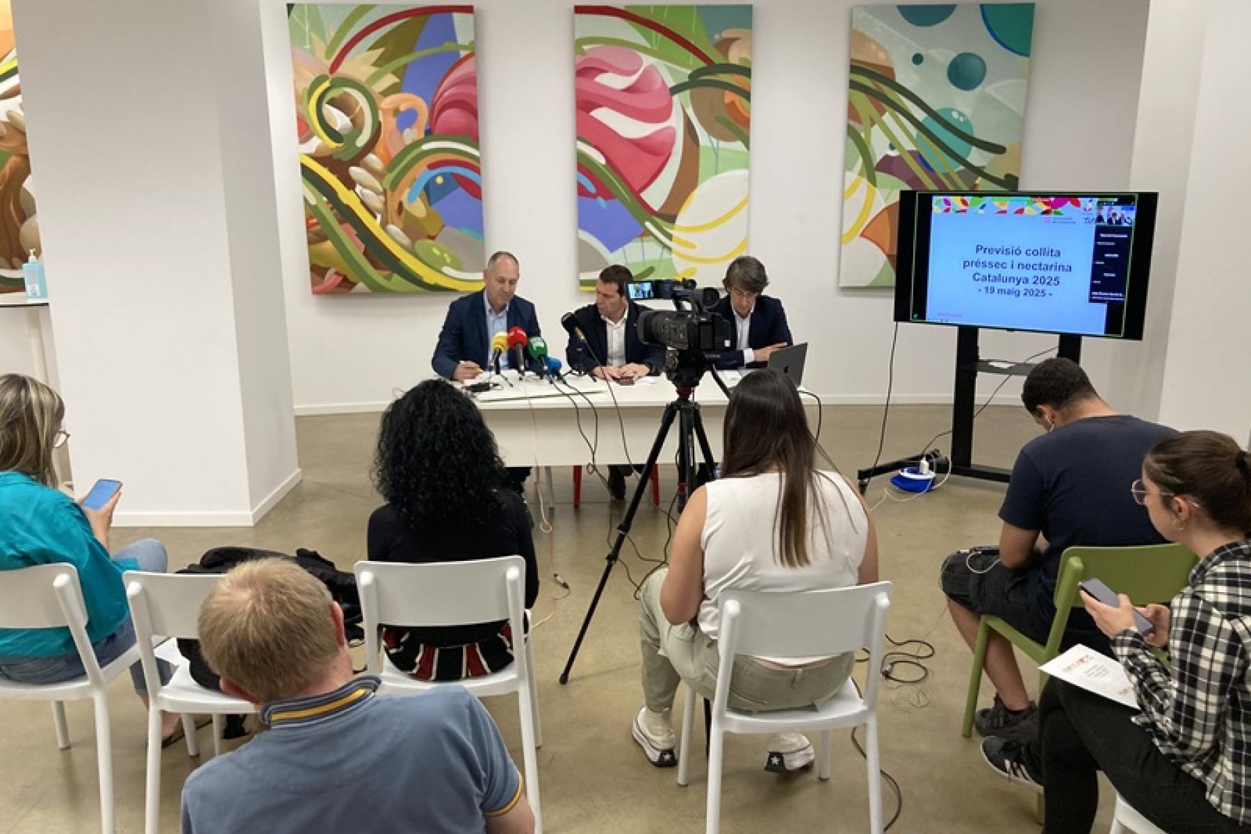 Pressekonferenz mit drei Personen an einem Tisch, davor sitzen einige Personen die zuhören und teilweise Notizen machen. Mikrofone und Kameras sind außerdem sichtbar.