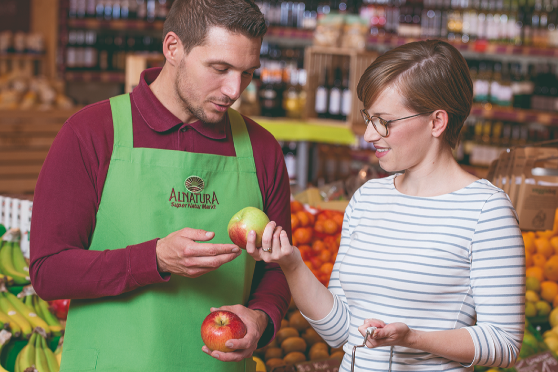 Mitarbeiter im Alnatura Super Natur Markt zeigt Kundin zwei Äpfel im Obst- und Gemüseregal