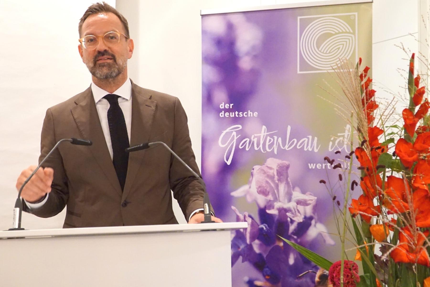 Bertram Fleischer steht hinter einem Rednerpult, neben einem Banner mit lila Blumen und Text 'der deutsche Gartenbau ist wert'