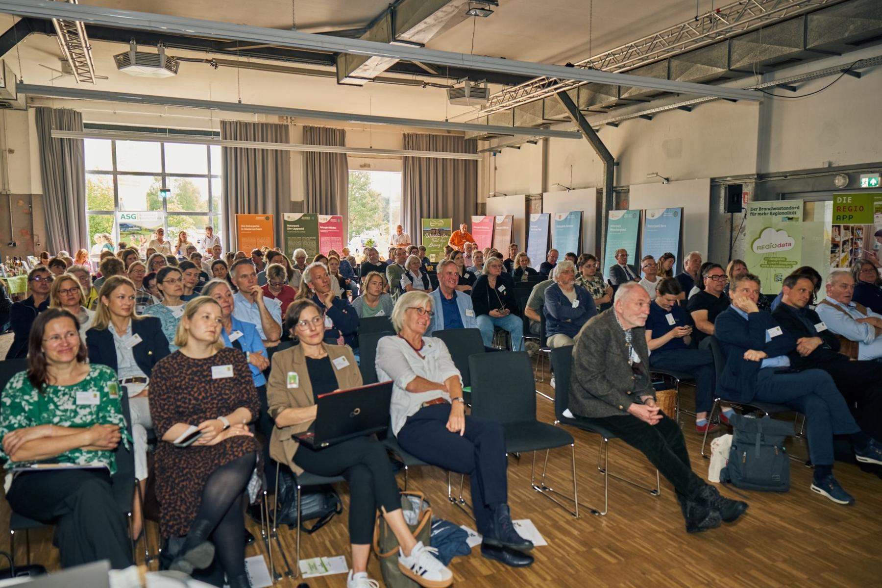 Große Gruppe von Menschen sitzt in einem hellen Konferenzraum und hört einem Vortrag zu, viele mit Namensschildern und Notizmaterialien