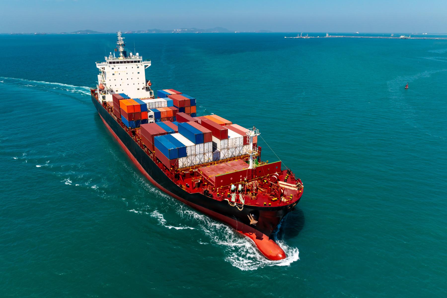 Containerschiff mit bunten Frachtcontainern fährt auf offenem Meer unter klarem Himmel, Küstenlinie im Hintergrund sichtbar