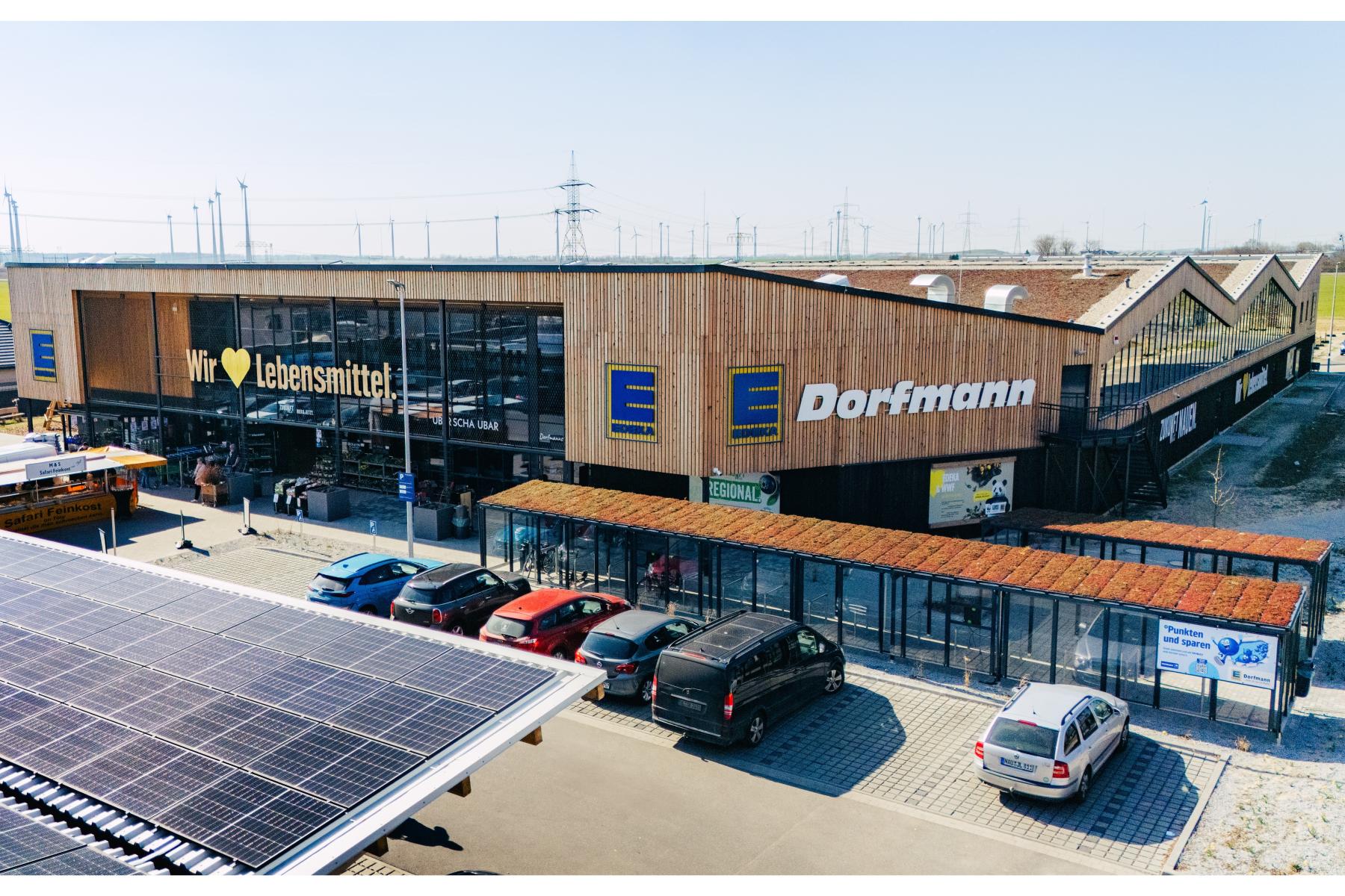 Edeka Dorfmann Supermarkt mit Holzfassade und großen Fenstern, mehrere Autos auf dem Parkplatz, sichtbare Solarpanels auf dem Dach.