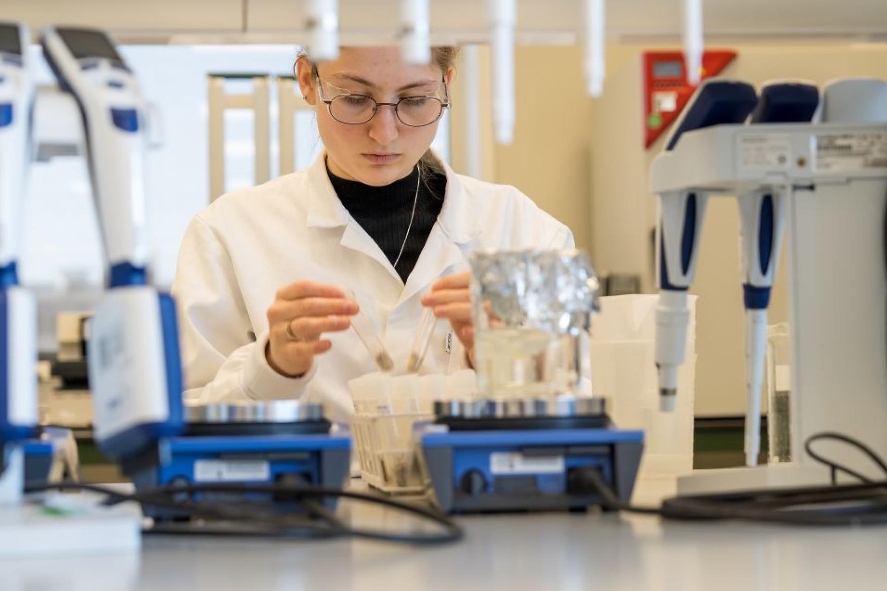 Person in weißem Laborkittel sitzt an einem Labortisch mit Laborgeräten und hält ein kleines Objekt in der Hand.