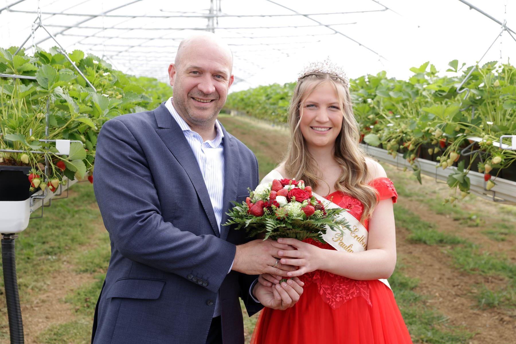 Landwirtschaftsminister Ingmar Jung und Erdbeerkönigin Paulina I. mit Krone und Schärpe, die einen Erdbeerstrauß hält, in einem Gewächshaus
