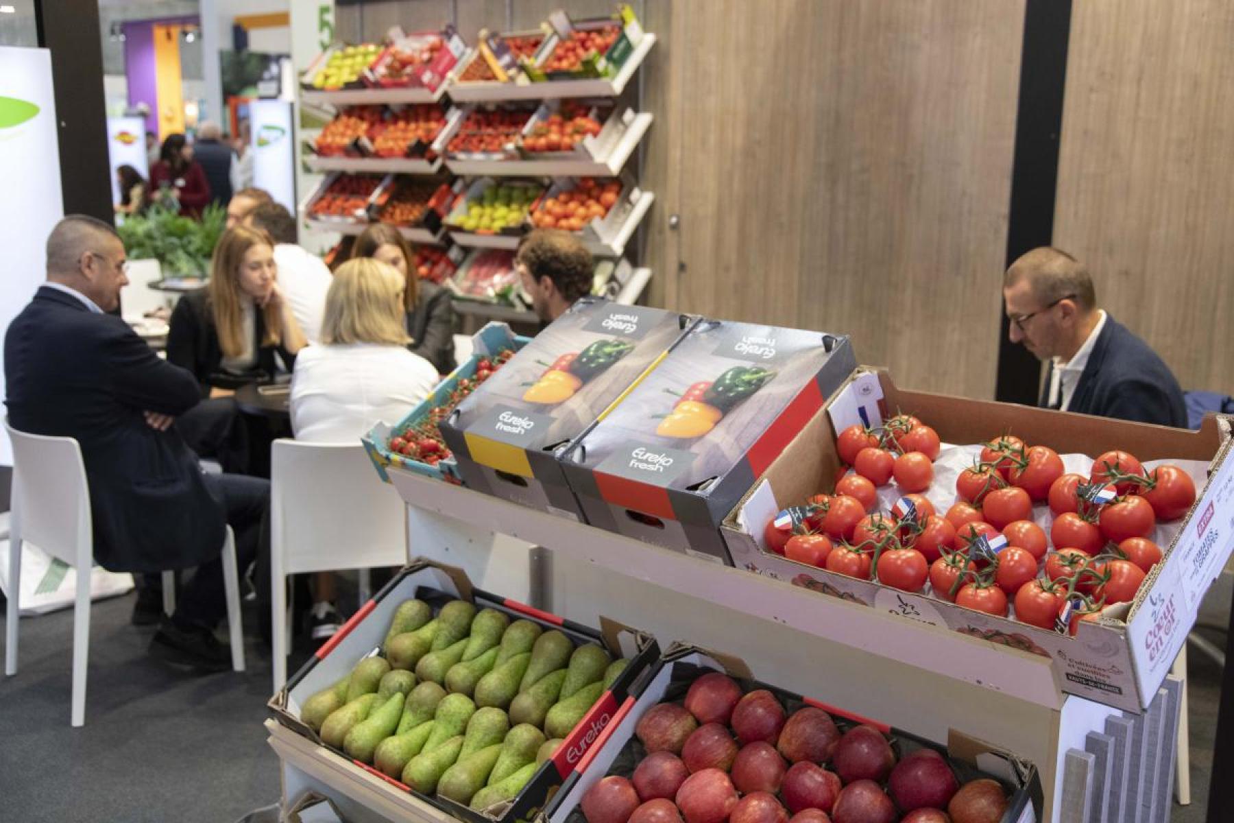 Mehrere Personen sitzen an einem Tisch in einem Messebereich, im Vordergrund Kisten mit Tomaten, Avocados und Granatäpfeln