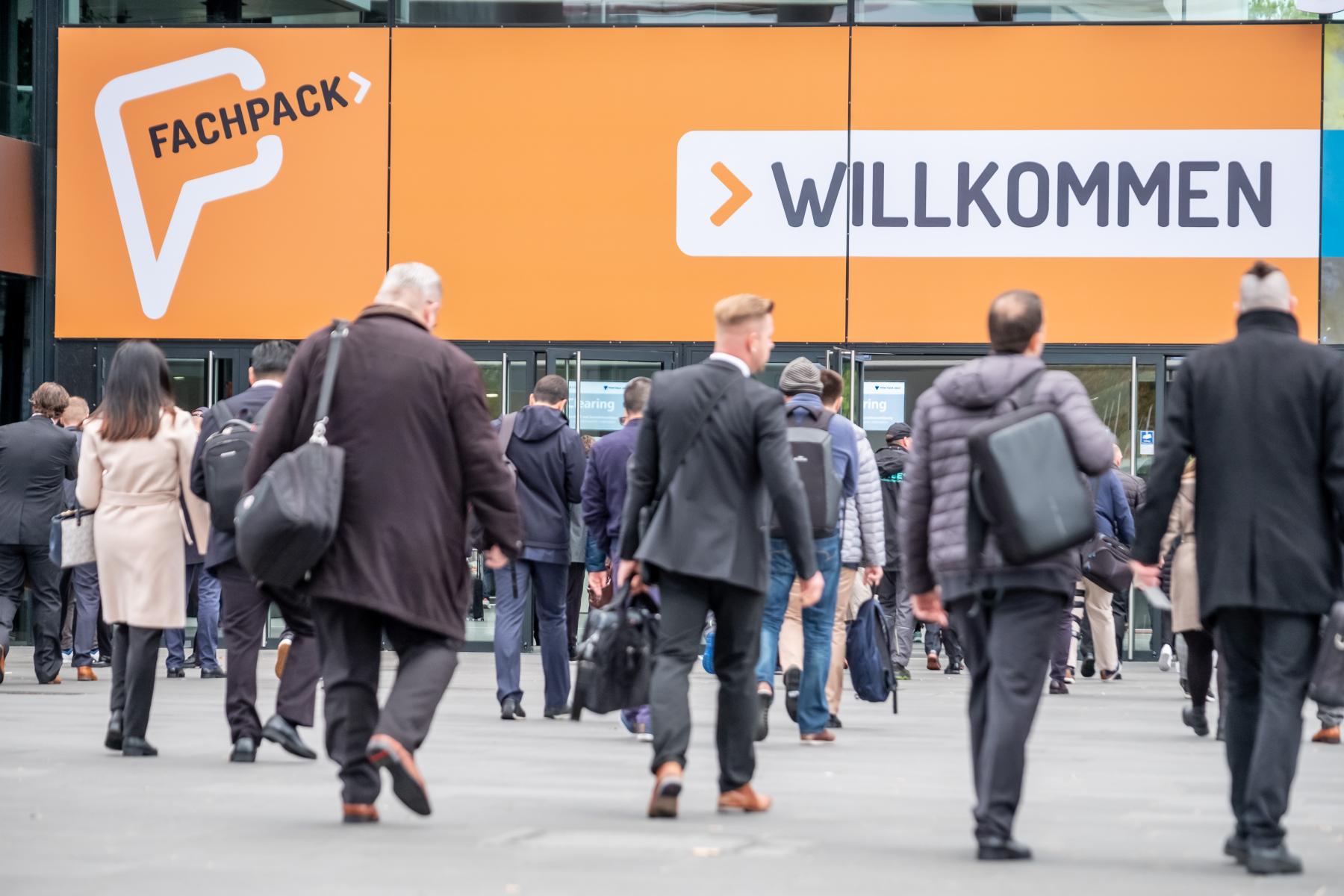 Menschen gehen auf den Eingang der Fachpack-Messe zu, großes orangefarbenes Schild mit 'Fachpack' und 'Willkommen'