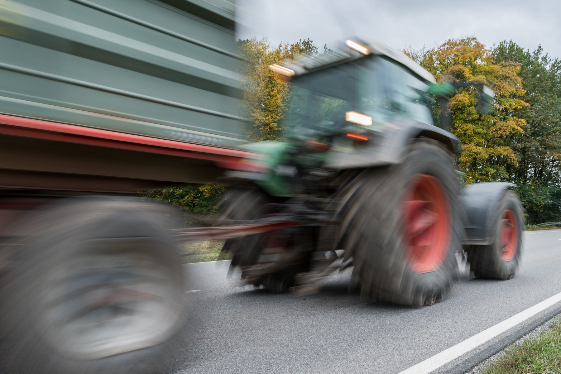Fahrender Traktor mit Anhänger auf Landstraße, unscharf durch Bewegung, Bäume im Hintergrund