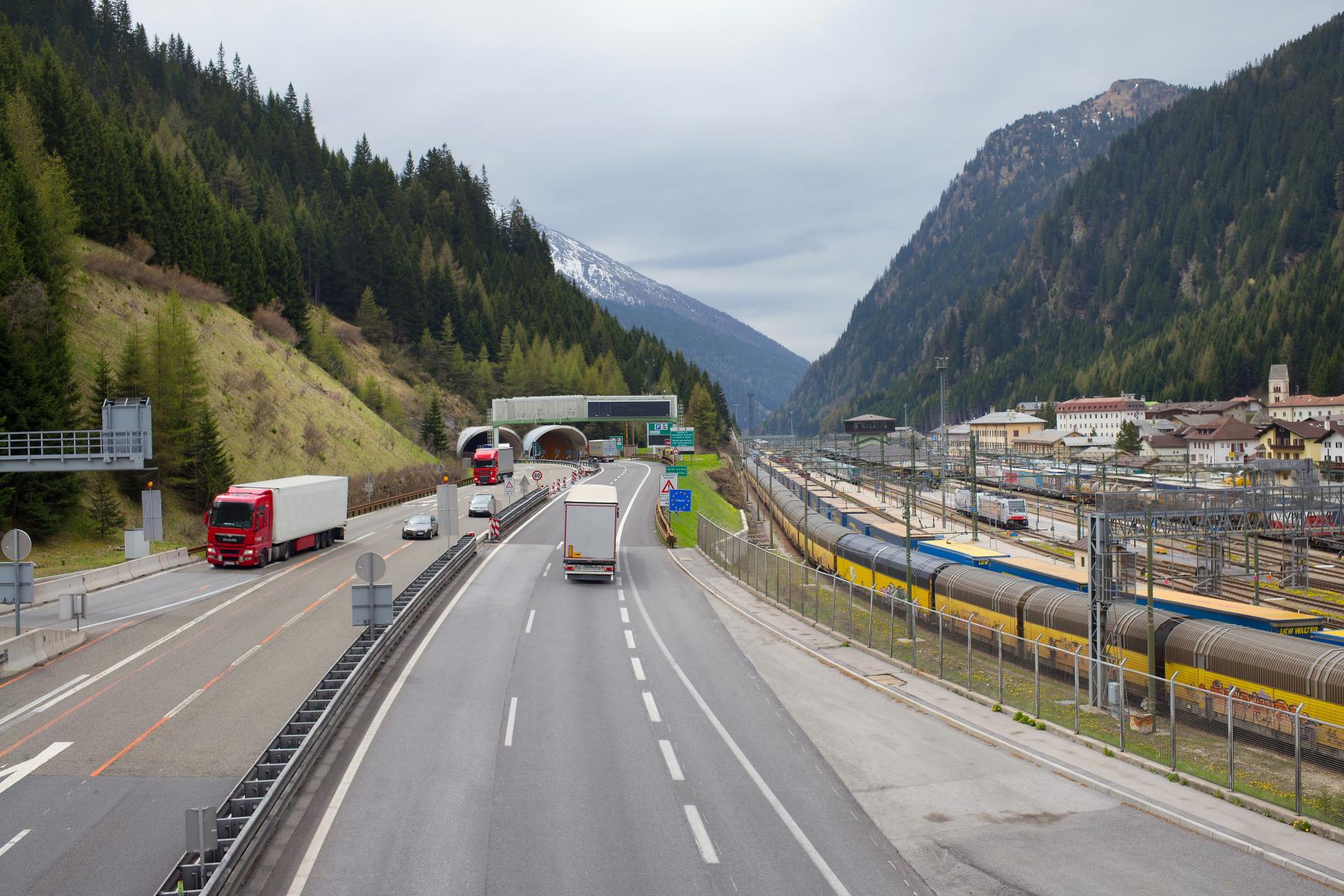 Autobahn mit Lastwagen und Autos am Brennerpass zwischen bewaldeten Bergen und einem Bahnhof mit Güterzügen
