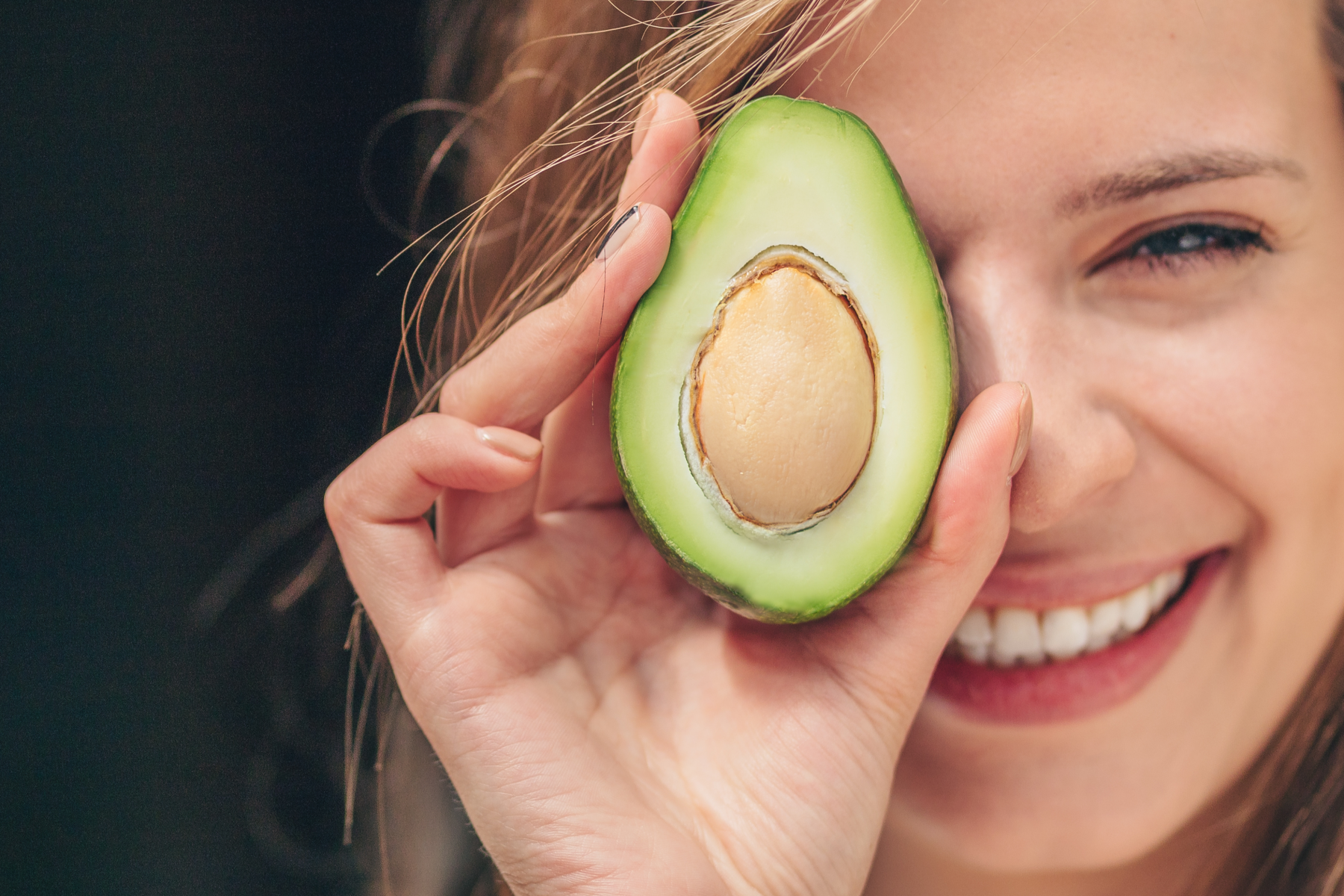 lächelnde Frau hält sich eine halbierte Avocado mit sichtbarem Kern vors Auge