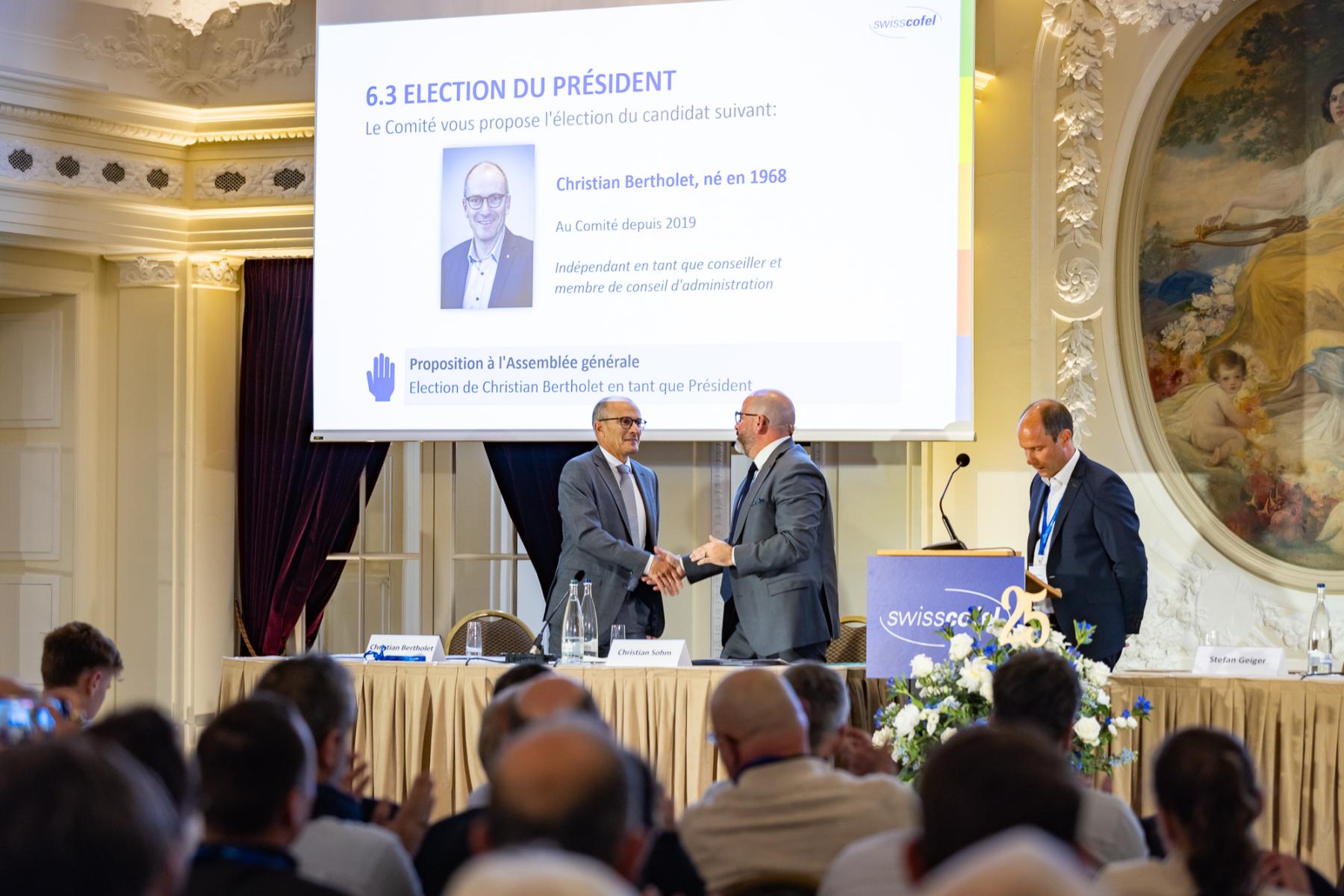 Christian Sohm gratuliert Christian Bertholet zur Wahl als Präsident, beide stehen, im Hintergrund ein Bildschirm mit Wahlinformationen.