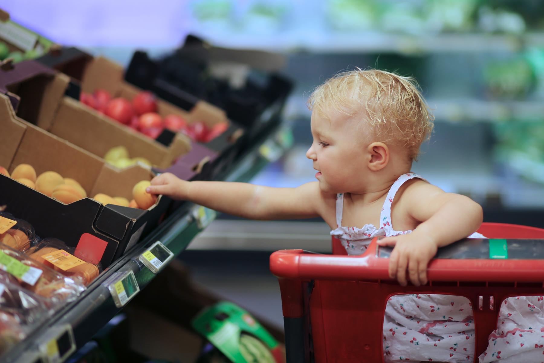 Kleinkind sitzt in einem Einkaufswagen und greift in die Obstauslage eines Supermarktes