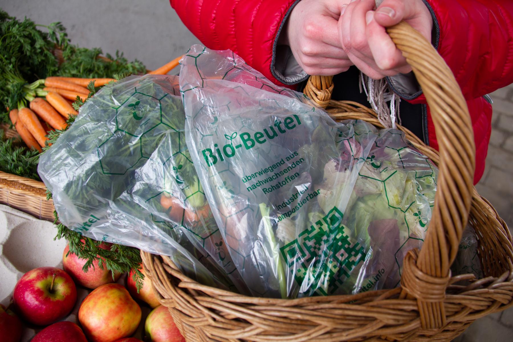 Hände halten Korb mit kompostierbaren Bio-Beuteln aus nachwachsenden Rohstoffen, umgeben von frischem Obst und Gemüse