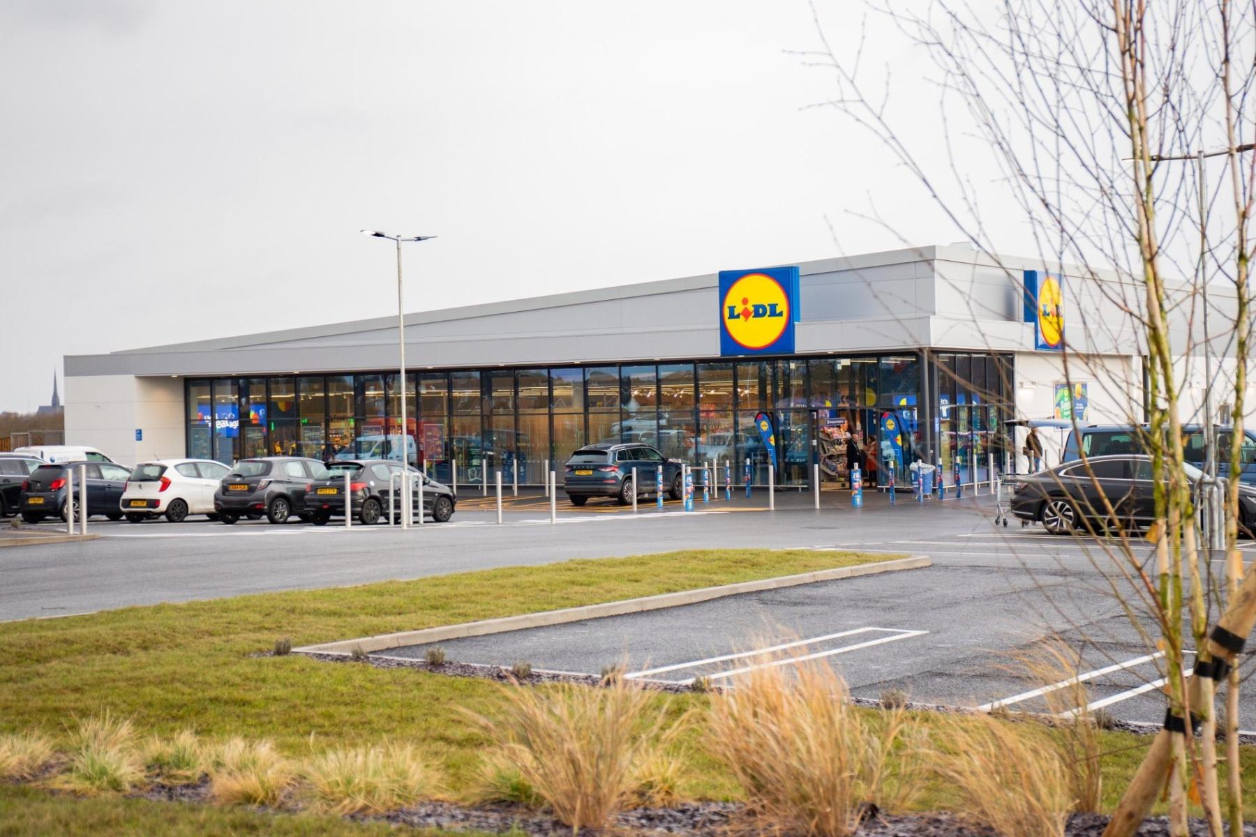 Lidl-Filiale mit Glasfassade, Parkplatz mit mehreren Autos und kahlen Bäumen im Vordergrund