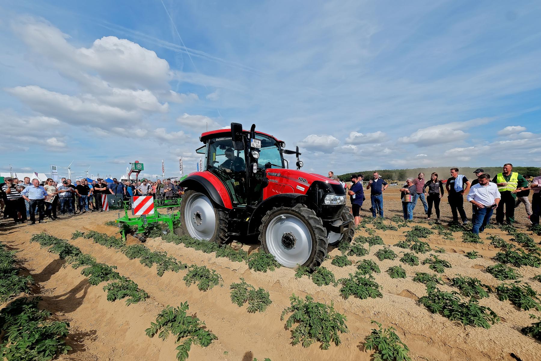 Roter Traktor bei Maschinenvorführung auf Kartoffelfeld, umgeben von Zuschauern unter blauem Himmel