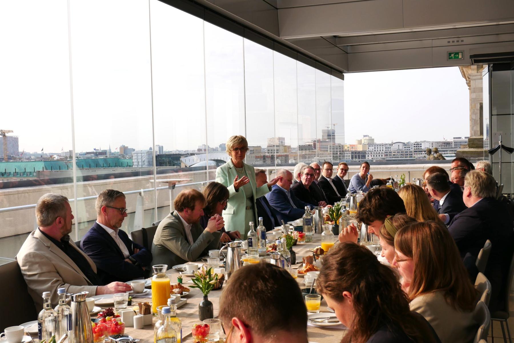 Menschen sitzen an einem langen Tisch auf einer Terrasse mit Blick auf eine Stadt, eine Frau steht und spricht zu der Gruppe während eines Frühstückstreffens.