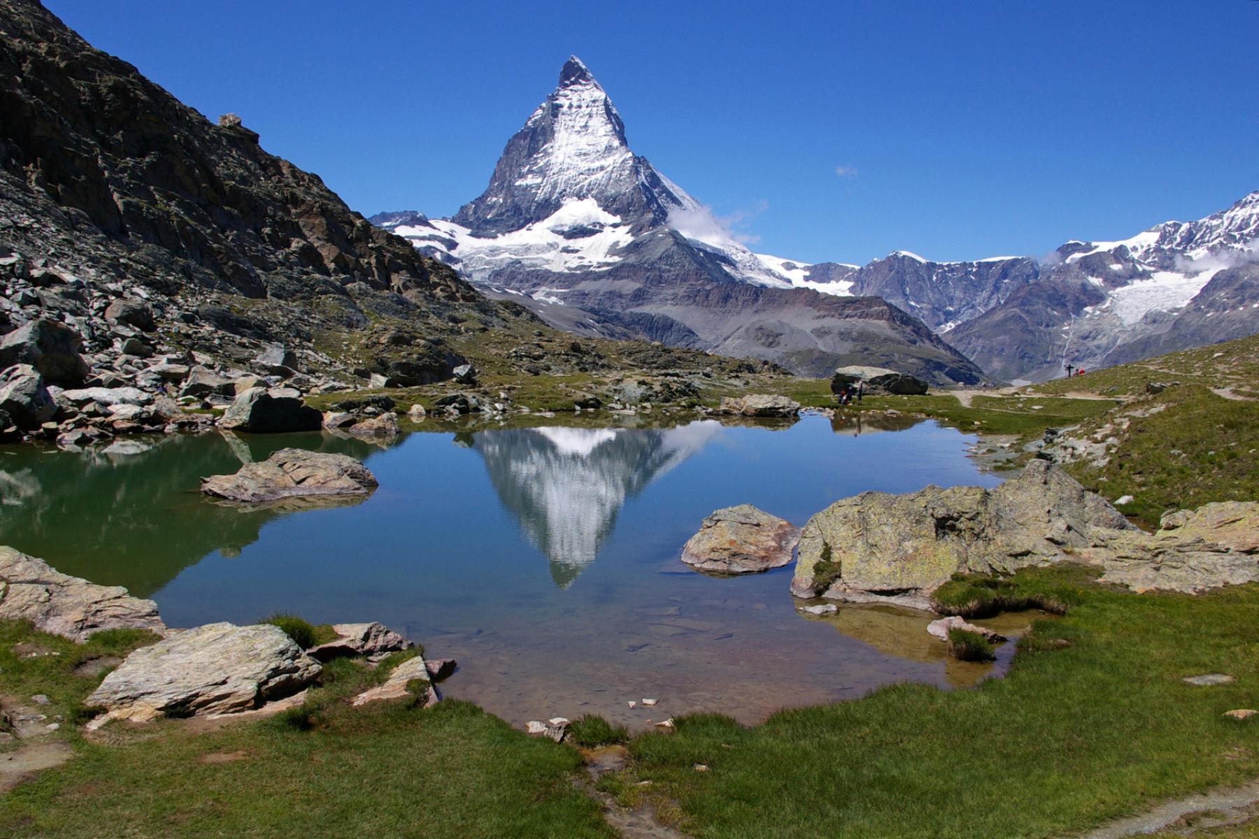 Matterhorn mit schneebedeckter Spitze, reflektiert in einem Bergsee, umgeben von Felsen und grüner Wiese