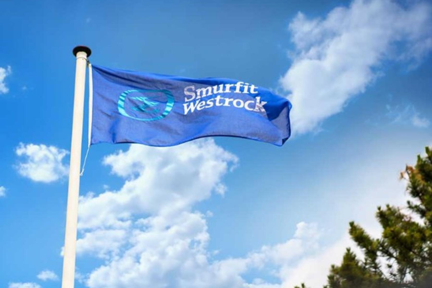 Blaue Flagge mit weißem Schriftzug 'Smurfit Westrock' weht an Fahnenmast vor blauem Himmel mit Wolken