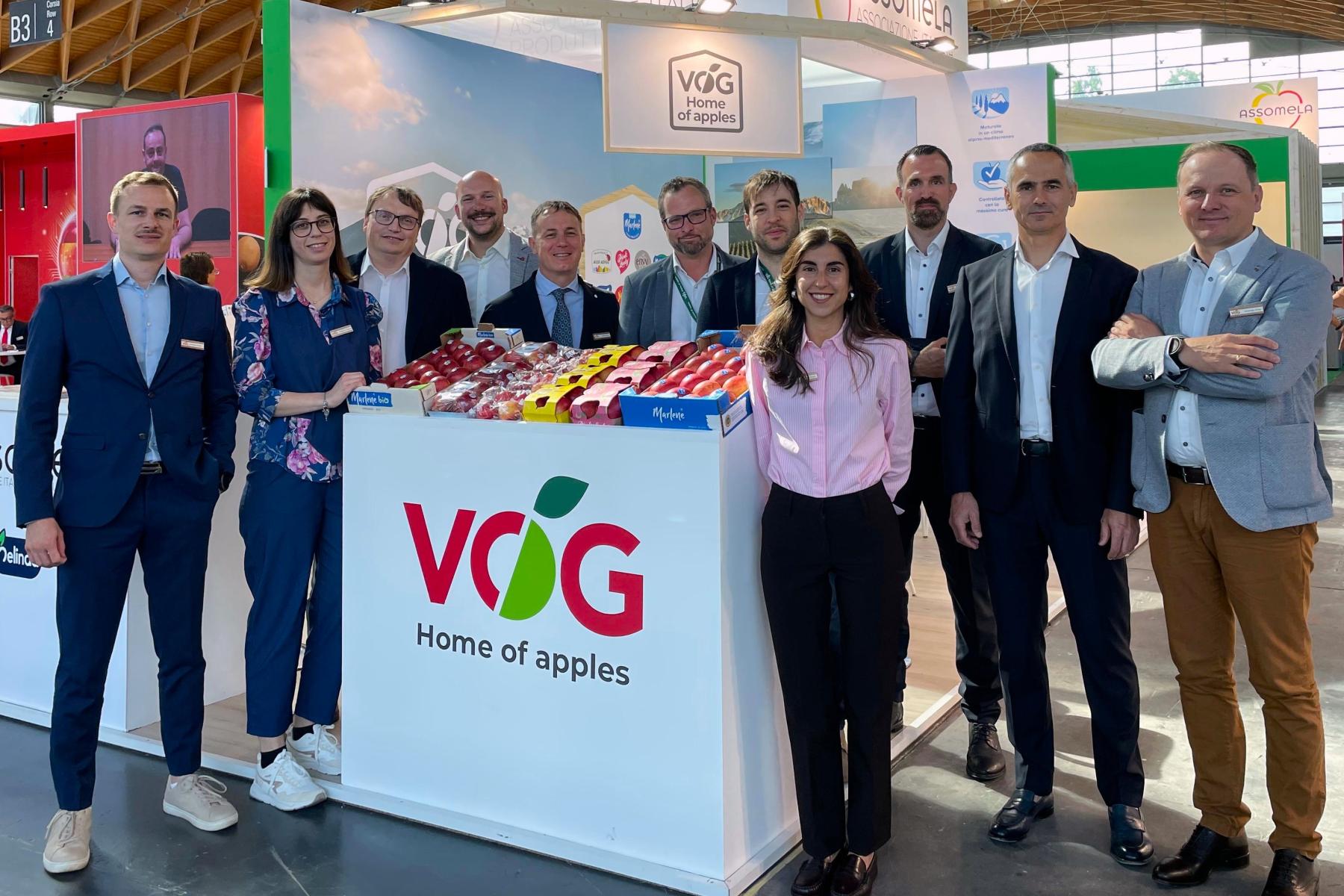 Das Team VOG steht hinter einem Messestand mit dem Logo 'VOG Home of apples' der mit verschiedenen Apfelsorten in Kisten dekoriert ist