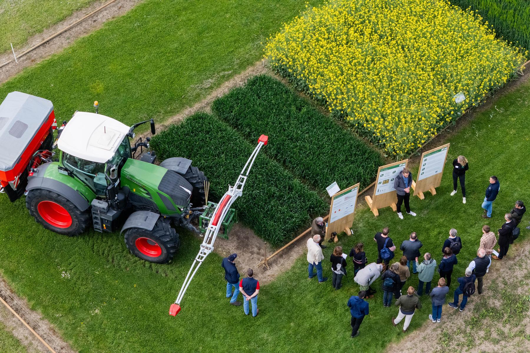 Gruppe von Menschen steht auf einem Feld neben einem grünen Traktor mit Anbaugerät und mehreren Parzellen mit verschiedenen Pflanzen