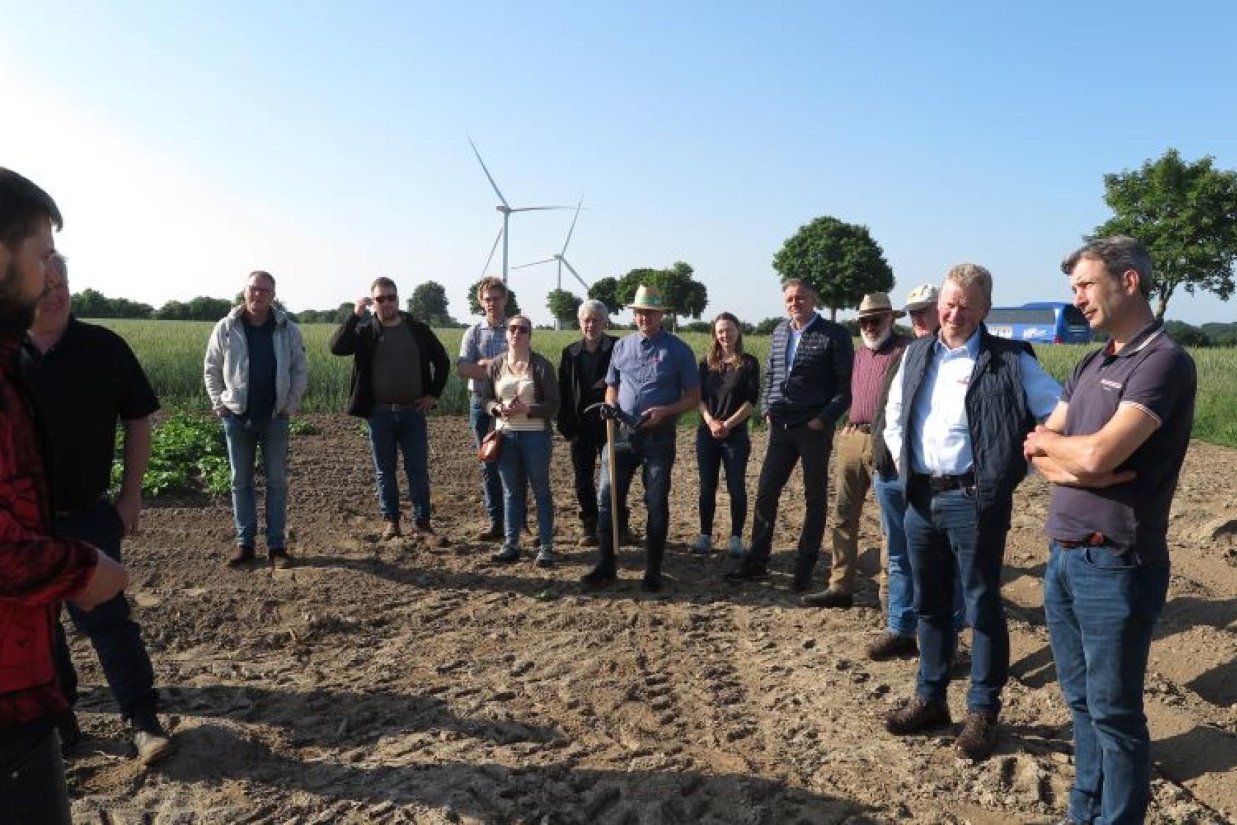 Gruppe von zwölf Personen steht auf einem Acker, im Hintergrund Windräder und Bäume unter blauem Himmel
