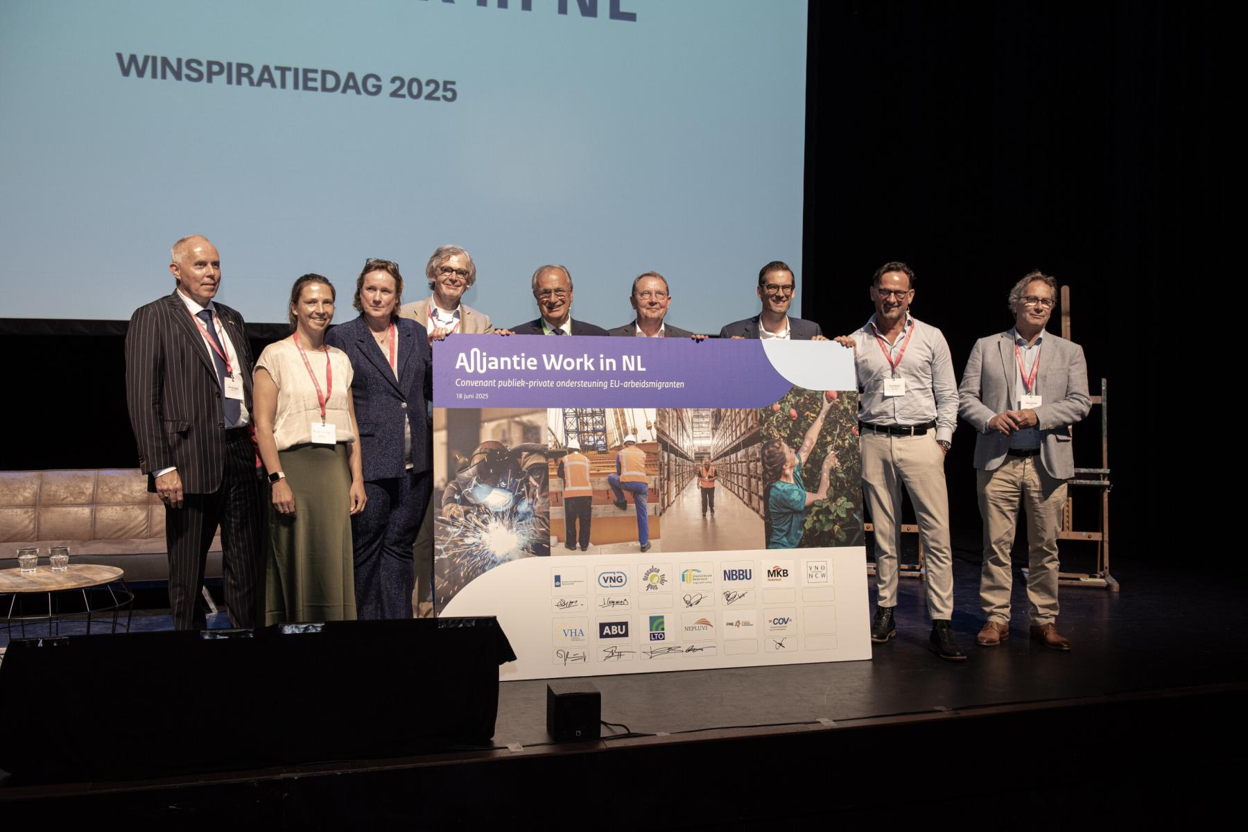 Gruppe von neun Personen auf Bühne halten großes Plakat mit dem Text 'Alliantie Work in NL' und Logos verschiedener Organisationen